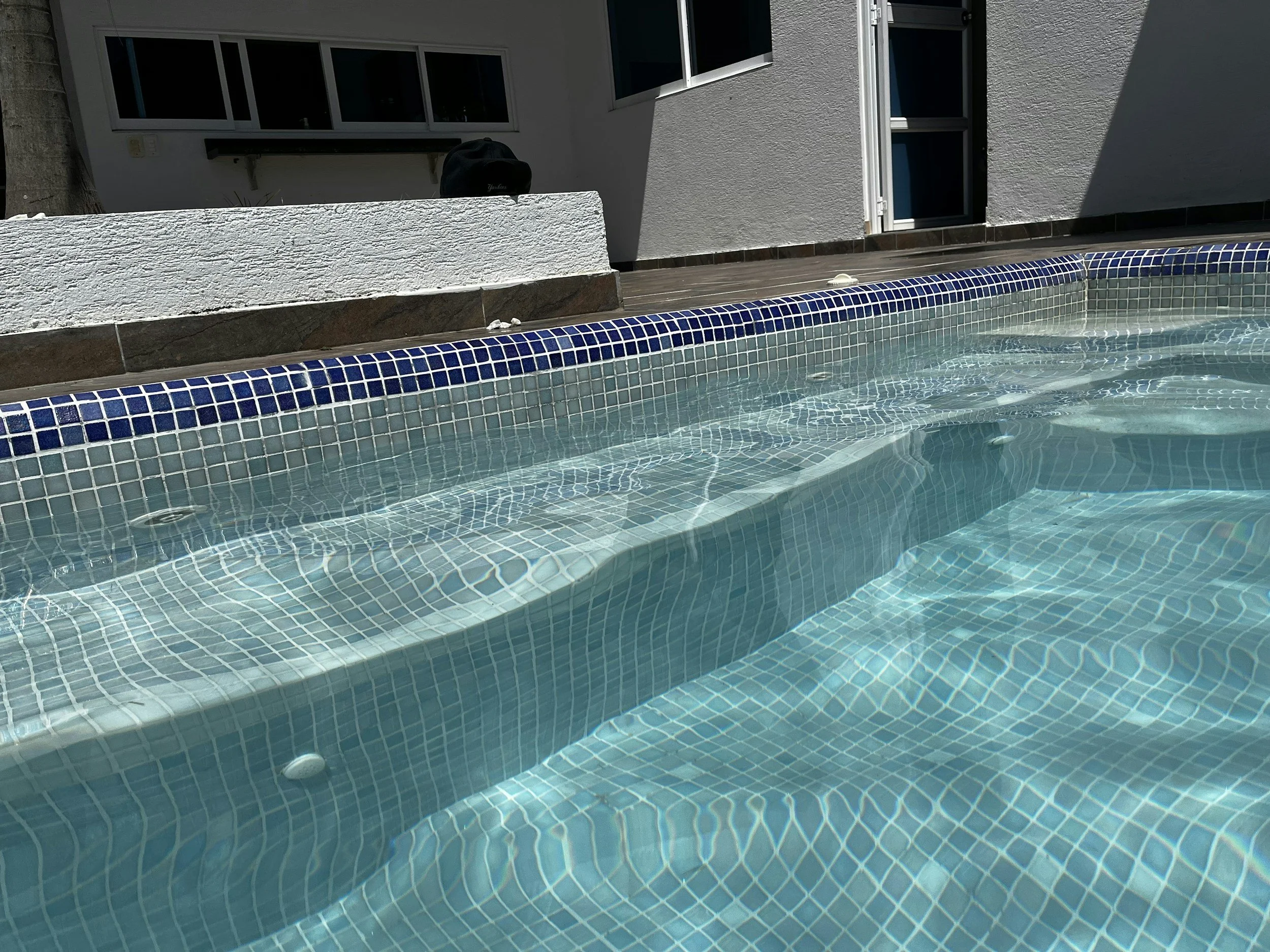 Swimming pool with clear water, blue tile border, and steps leading into the pool, surrounded by a wooden deck and adjacent to a white building with a door and windows.