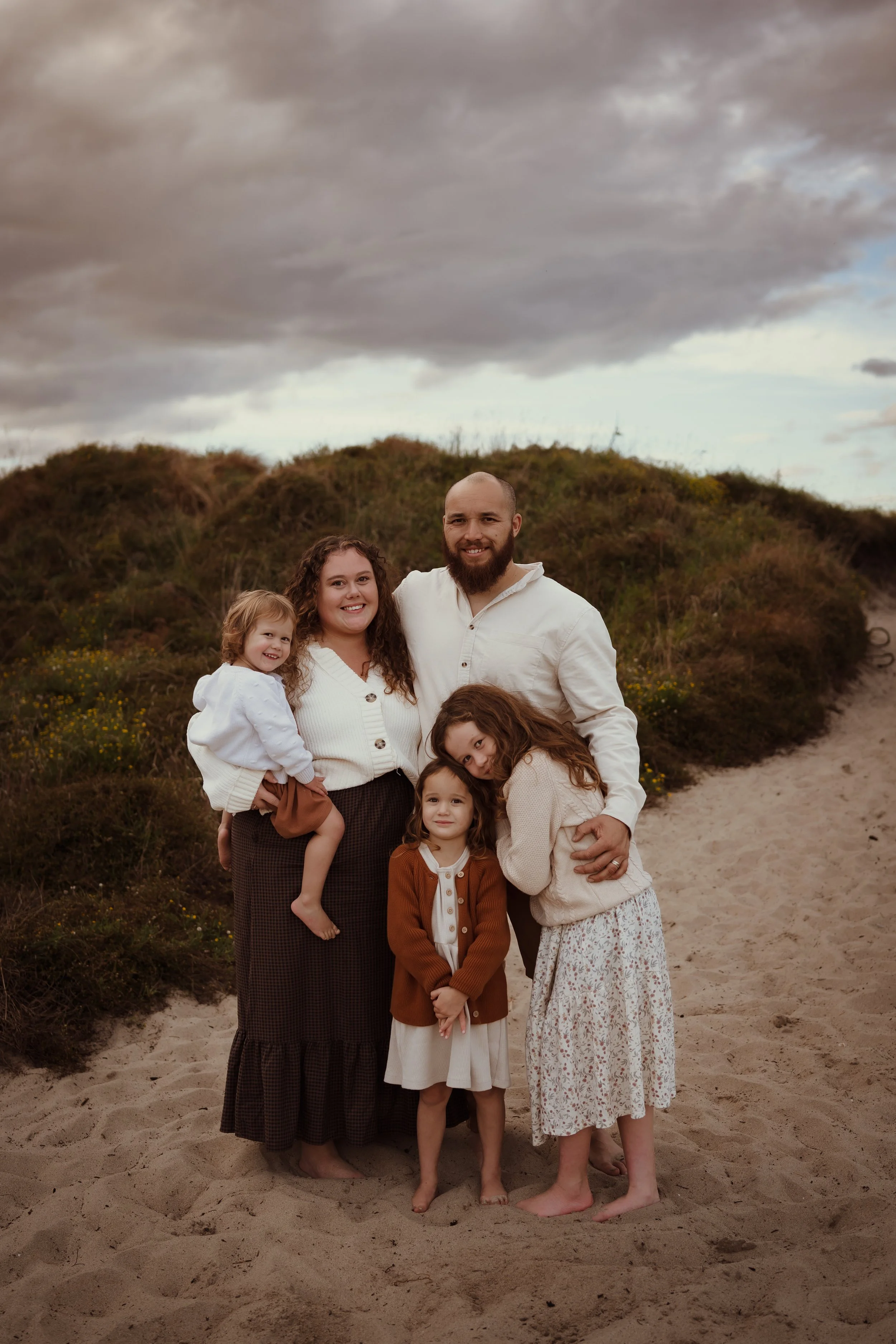 A family of six standing on a sandy Papamoa beach with dunes and a cloudy sky in the background in Tauranga. Shoreline Pool Services