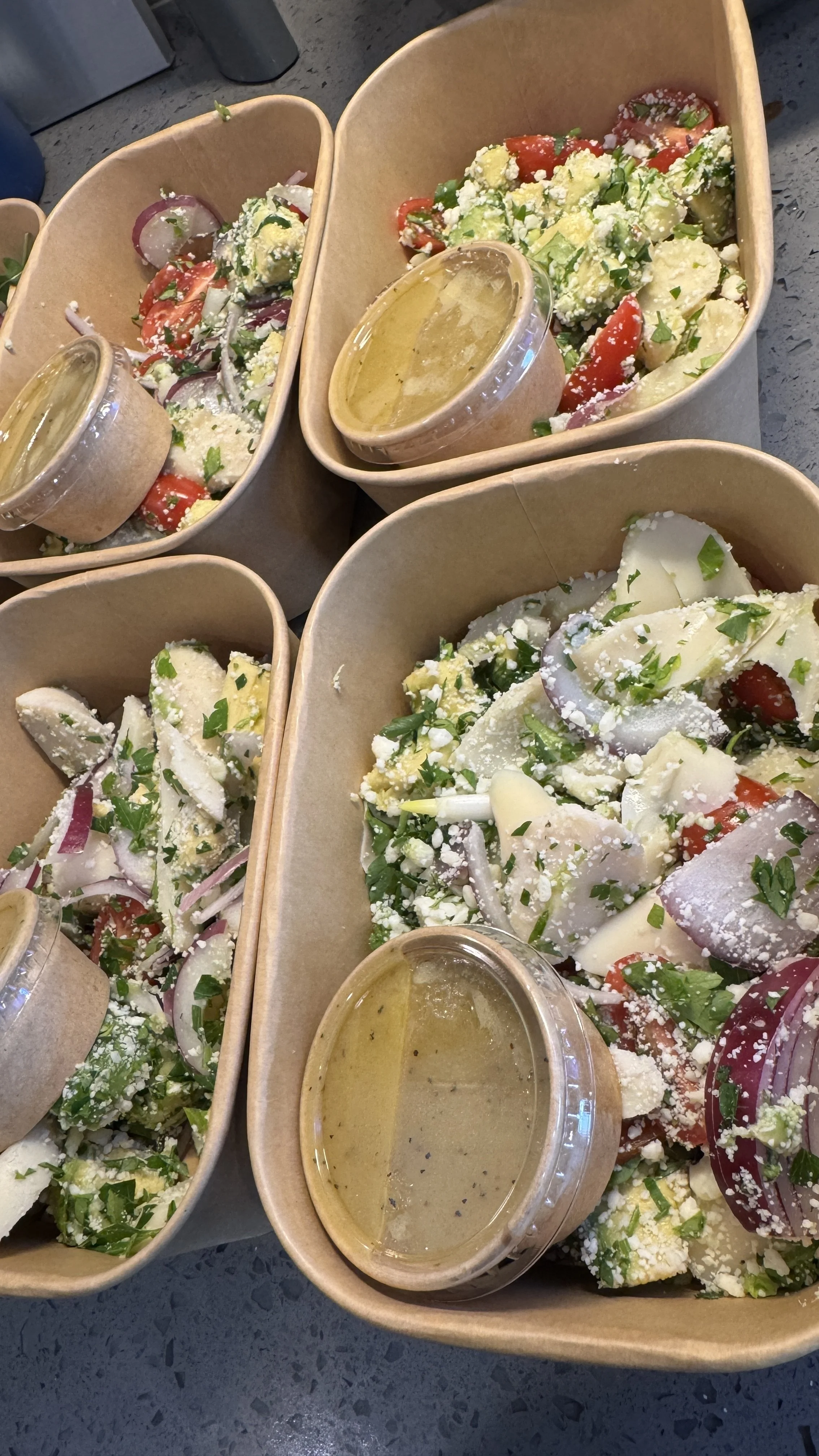Four containers of Greek salad with tomatoes, cucumbers, onions, and feta cheese, each accompanied by a small cup of dressing on a gray surface.