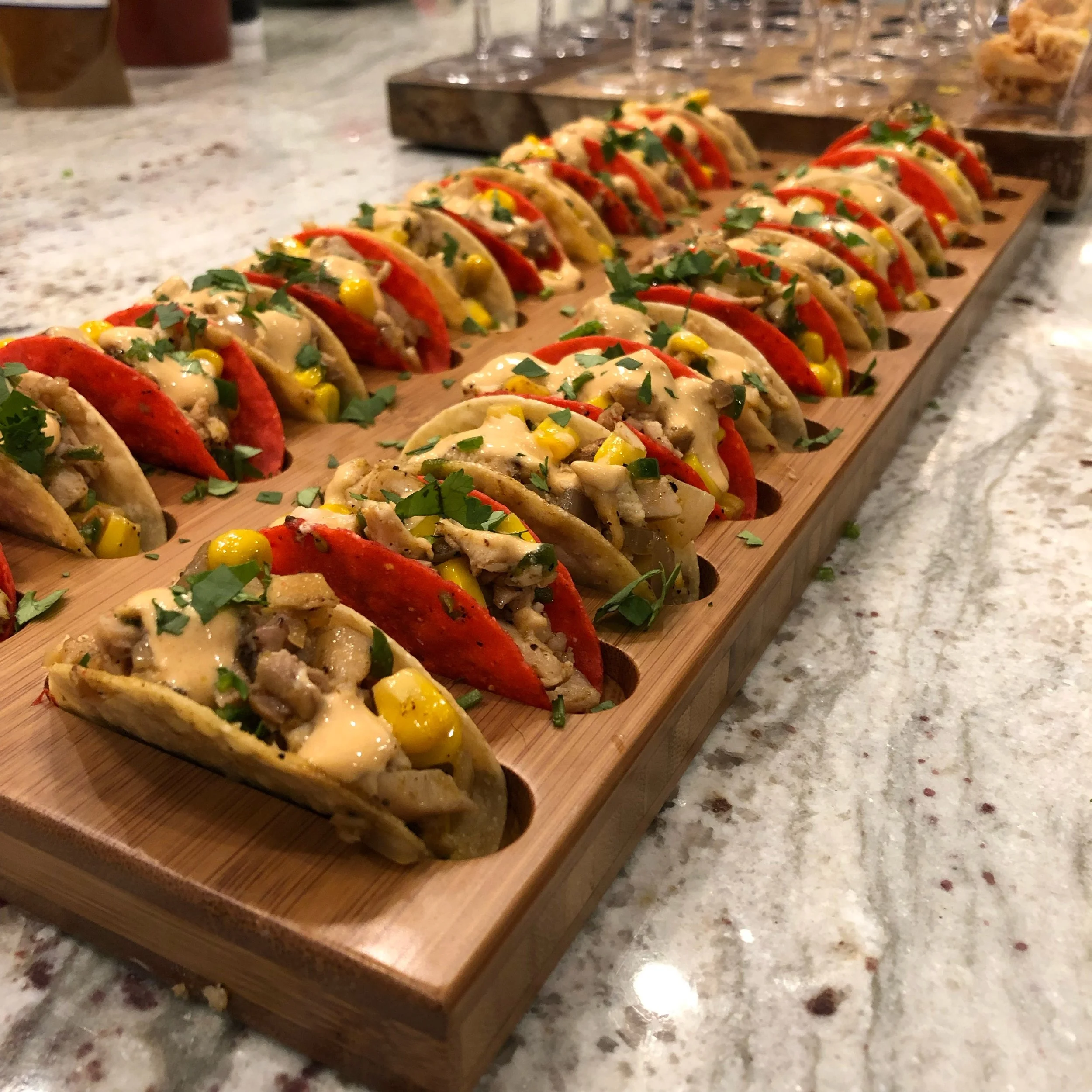A wooden serving board with multiple mini tacos filled with meat, corn, and herbs, garnished with chopped cilantro, arranged in a neat row on a marble countertop.