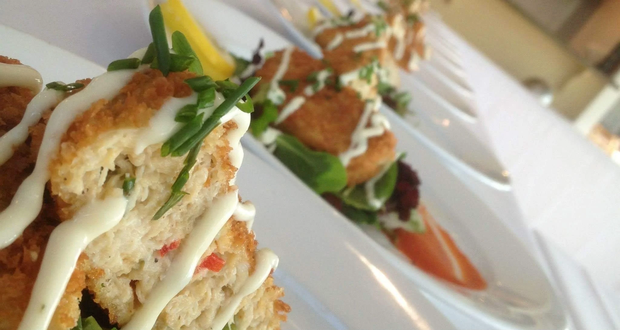 Close-up of crispy croquette topped with a white sauce and chopped green herbs, with another dish of fried food and salad in the background.