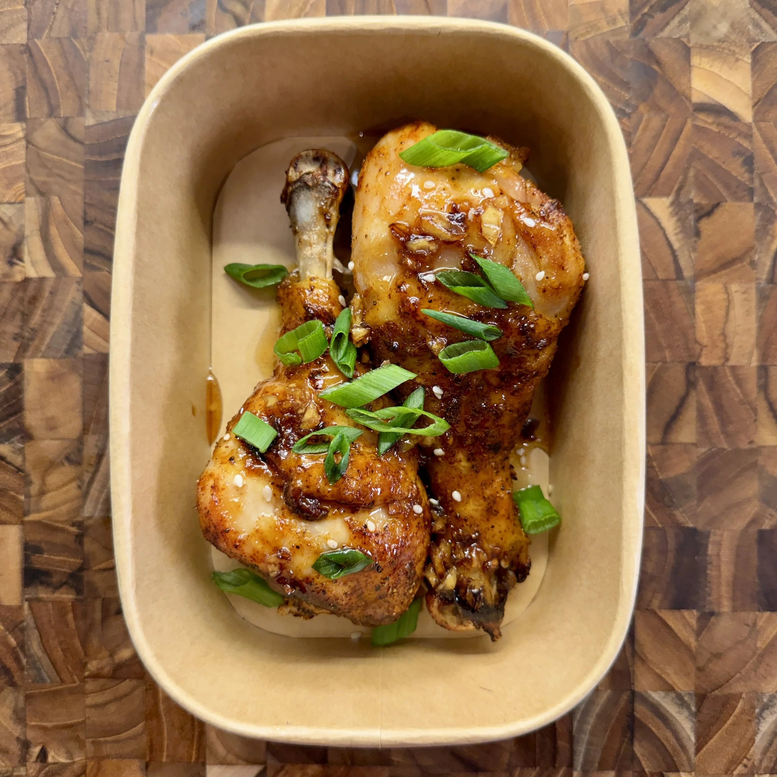 Baked chicken drumsticks garnished with chopped green scallions in a disposable paper tray.