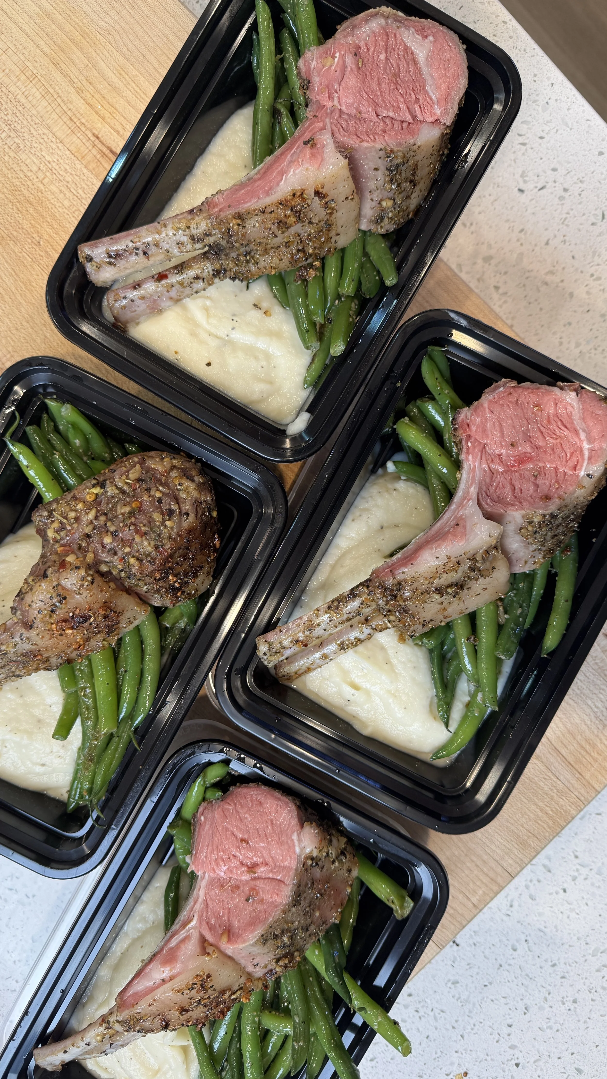 Three meal containers each with green beans, mashed potatoes, and a cooked lamb chop, with one container showing a seasoned, grilled lamb chop and another showing a plain cooked lamb chop.