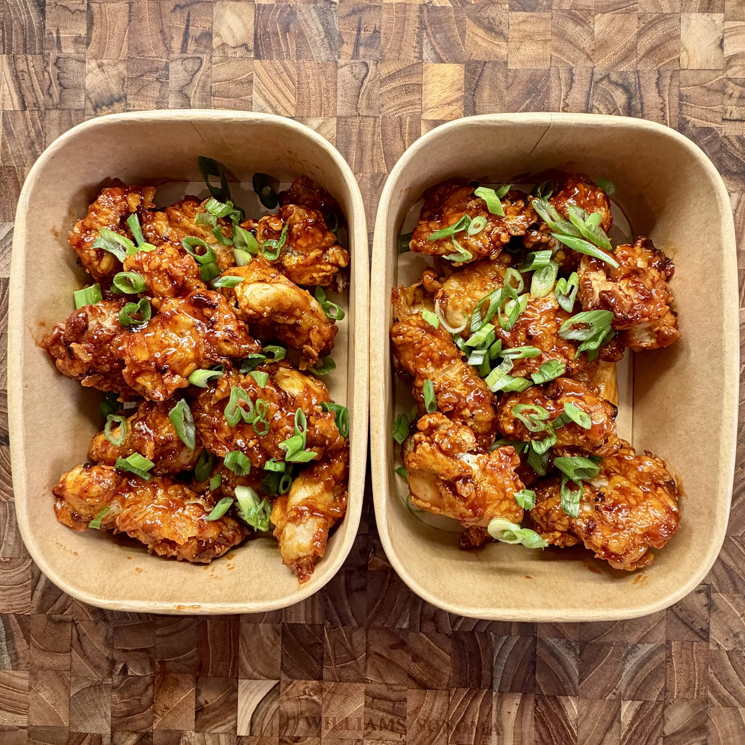 Two paper trays filled with spicy chicken wings garnished with chopped green onions, placed on a wooden surface.