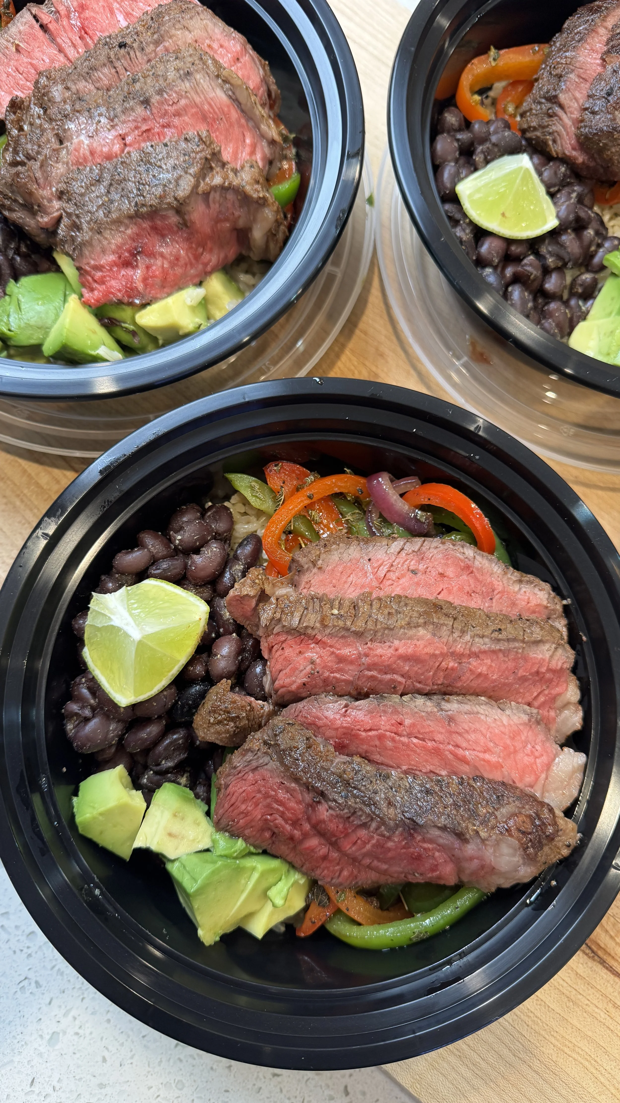 Three meal bowls with sliced cooked steak, black beans, avocado, lime wedge, and sautéed vegetables including peppers and onions.