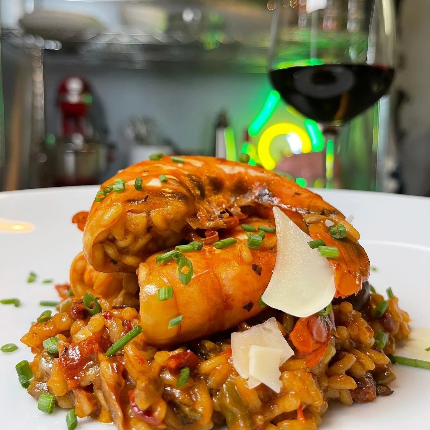 Close-up of a gourmet dish featuring a large stuffed mushroom on a bed of rice with vegetables, garnished with chopped chives and parmesan cheese, with a glass of red wine in the background.