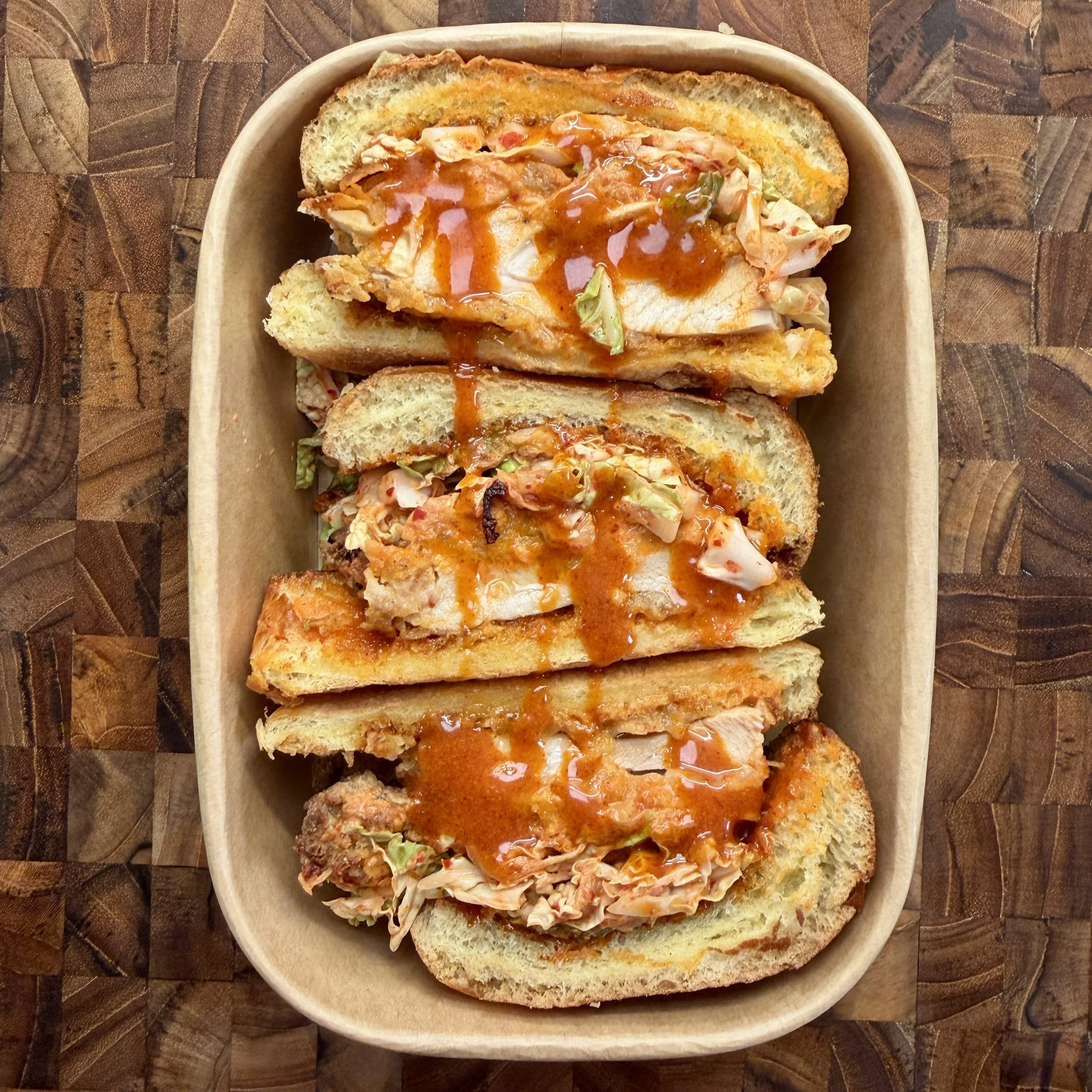 Four pulled pork sandwiches with barbecue sauce on toasted buns in a brown paper tray on a wooden surface.