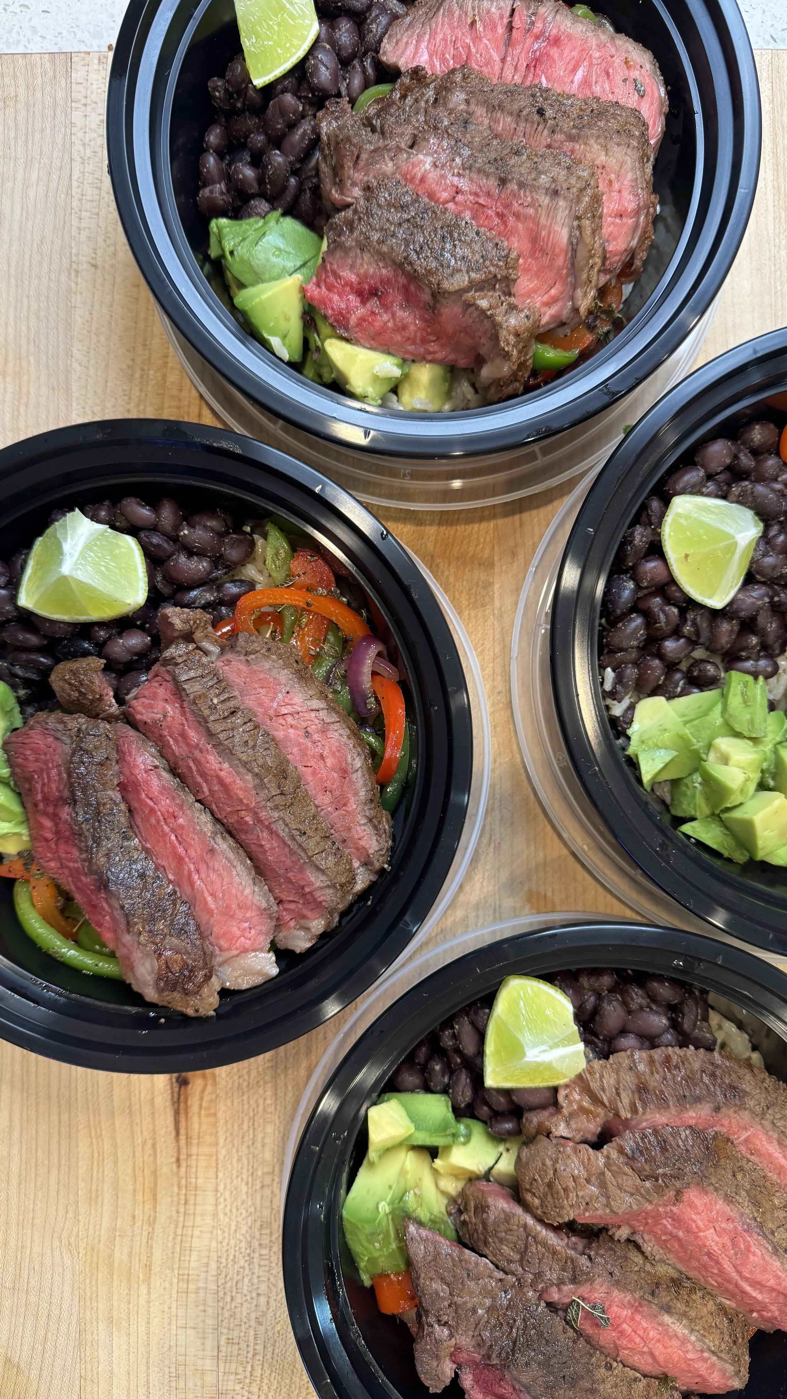 Four meal prep containers with sliced steak, black beans, diced avocado, lime wedges, and sautéed bell peppers and onions, placed on a wooden surface.