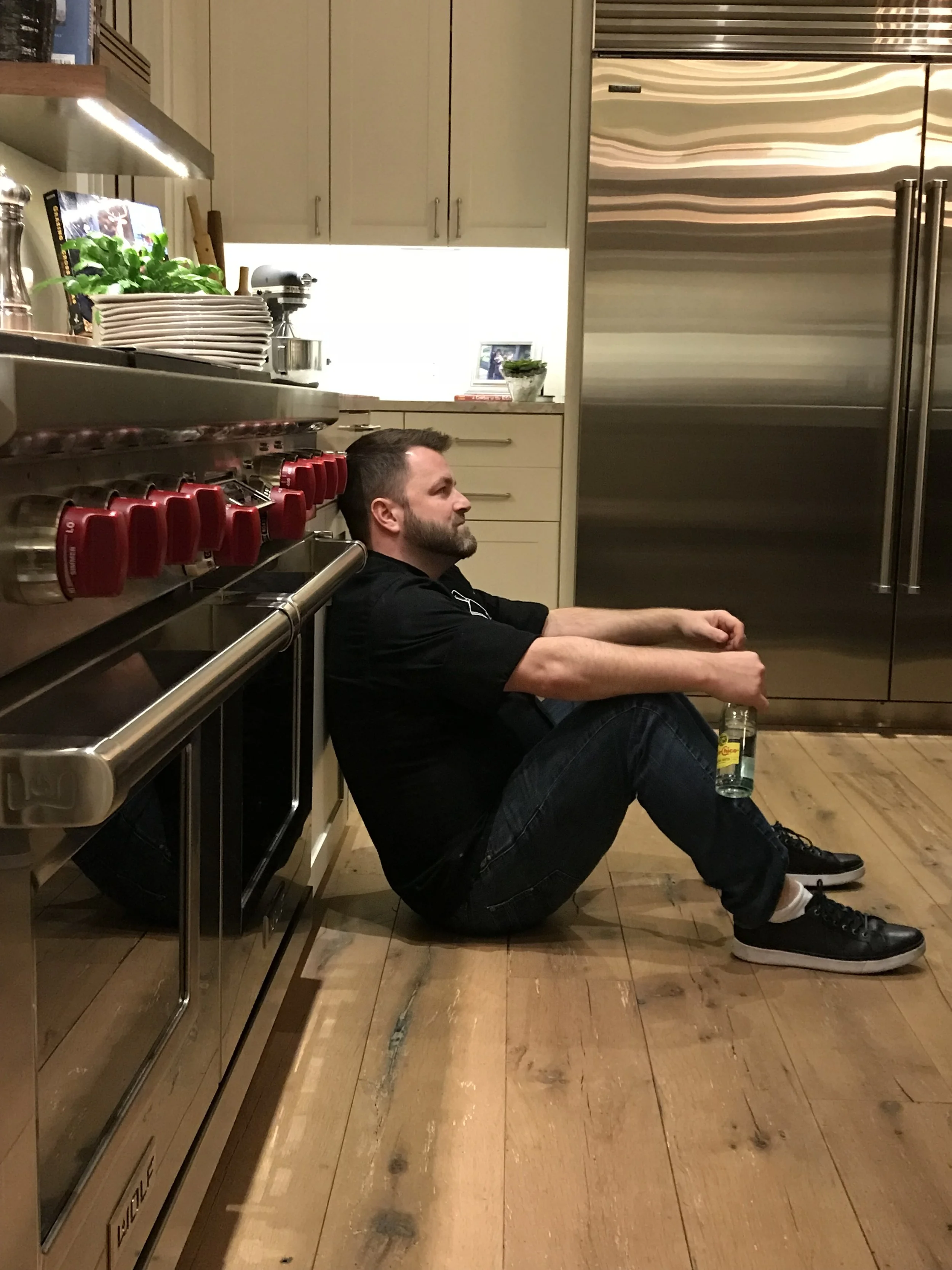 A man sitting on the kitchen floor, leaning against the cabinets near a stainless steel refrigerator, holding a bottled drink.