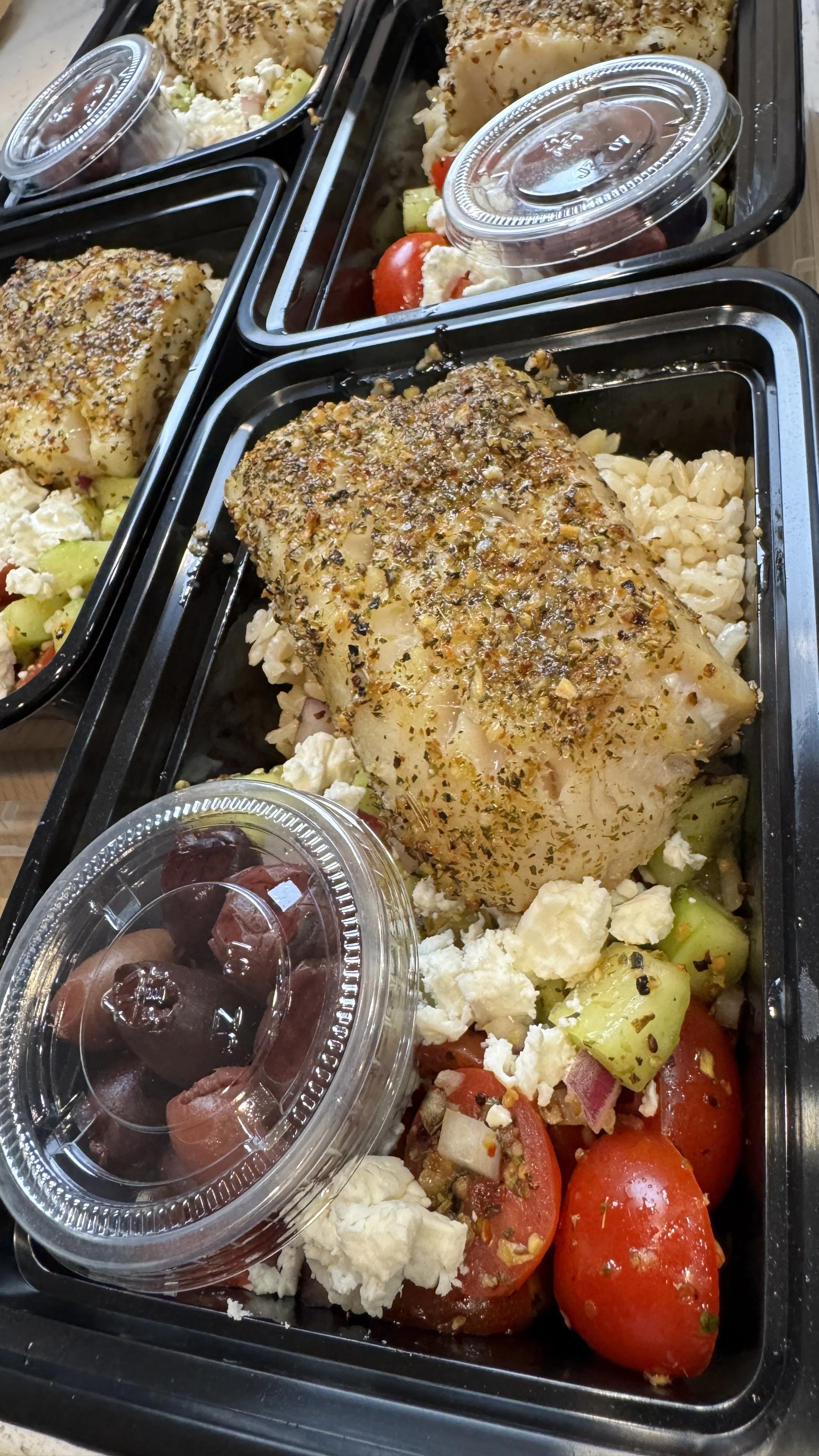 Baked seasoned fish fillet served with rice, tomato, cucumber, and feta cheese, accompanied by a small container of black olives.