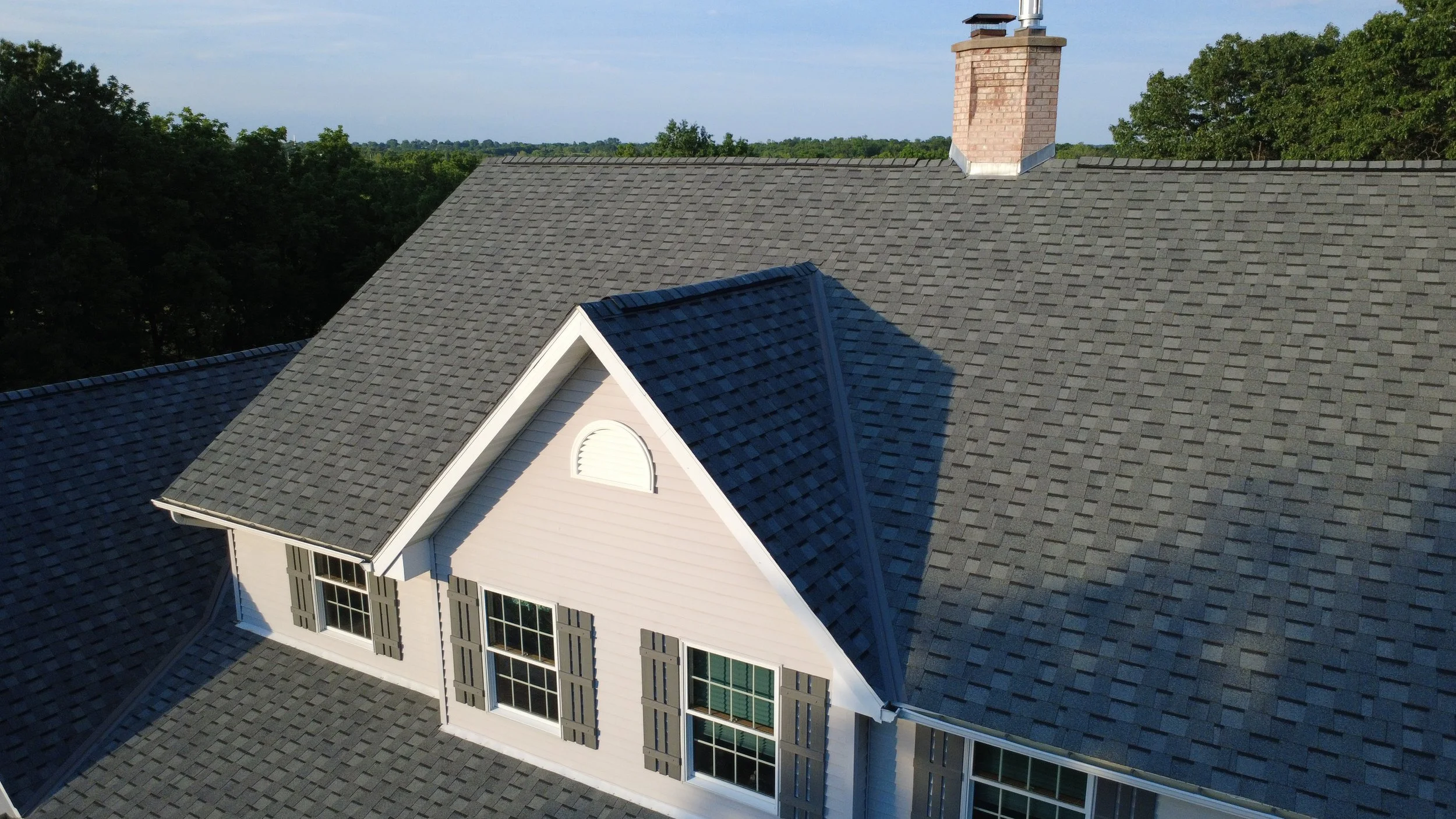 Aerial view of a house roof with gray shingles, dormer window, and brick chimney, surrounded by green trees and open land under a cloudy sky.