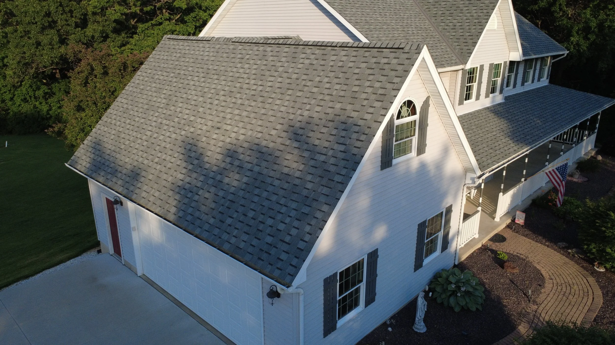 Aerial view of a white, two-story house with a gray gabled roof, multiple windows with black shutters, a front porch with white railings, and a red garage door. The house is surrounded by green trees and a landscaped yard.