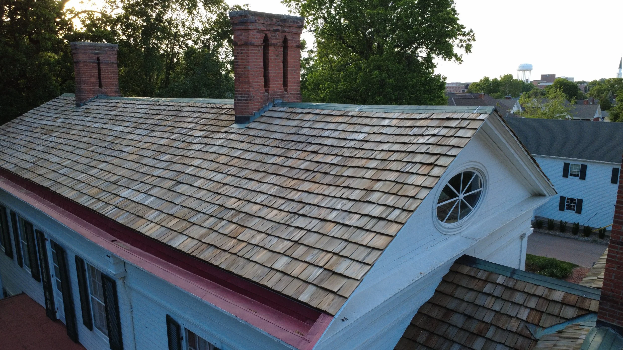 A rooftop with new wooden shingles, several brick chimneys, and a round window in a white house, with trees and other houses in the background.
