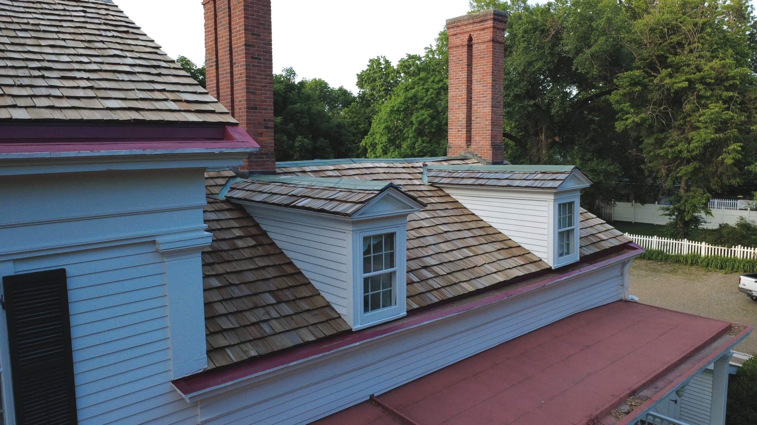 Aerial view of a house with multiple chimney stacks, dormer windows, shingled roof, and white wooden siding, surrounded by trees.