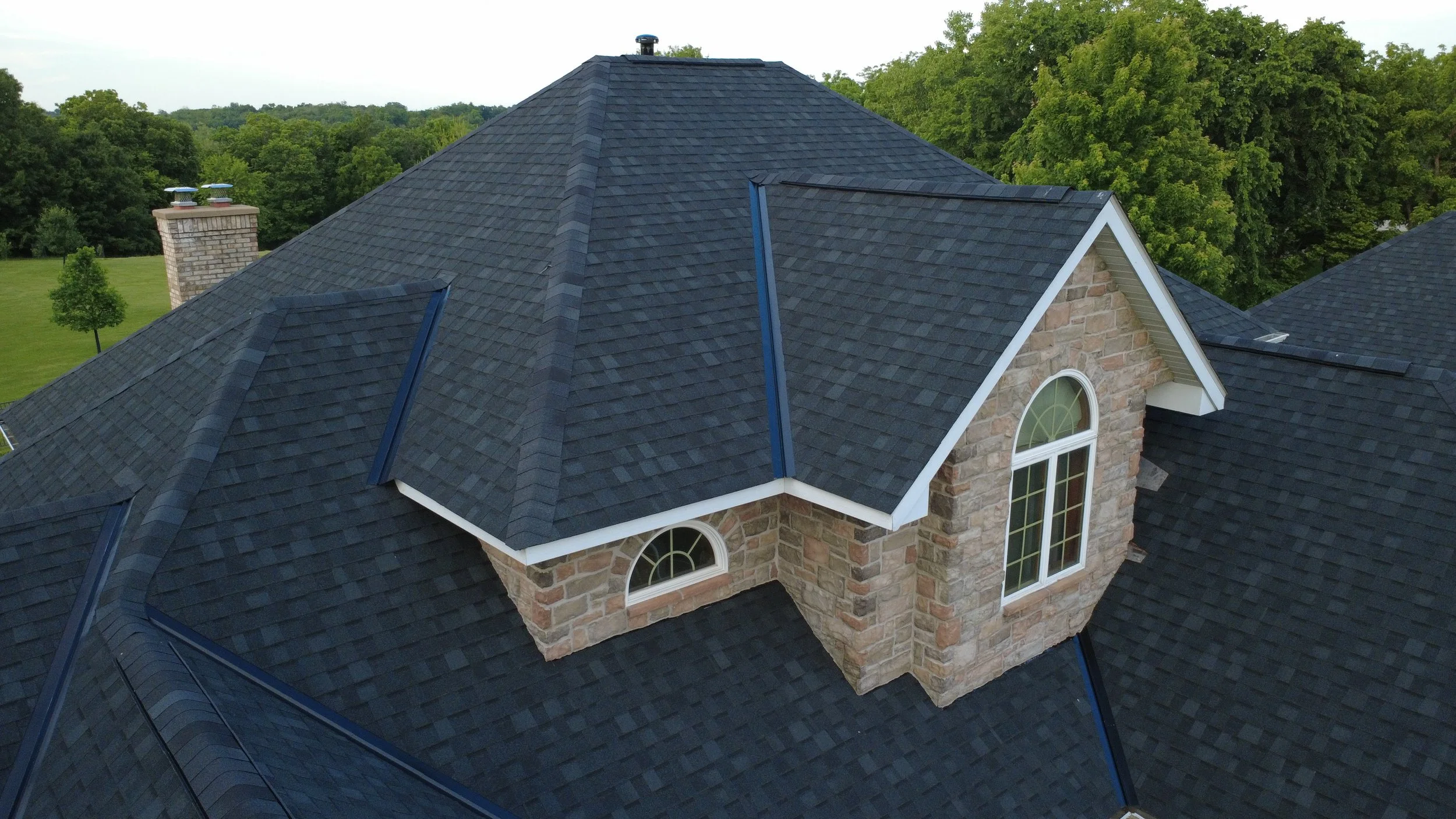 Aerial view of a house with dark shingles, brick walls, and large windows, surrounded by green trees and lawn.