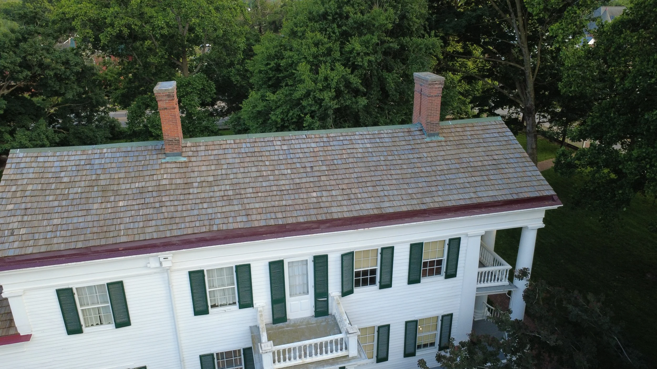 An aerial view of a house with a brown shingle roof, two brick chimneys, white exterior walls, dark green shutters, and a porch. The house is surrounded by tall green trees.