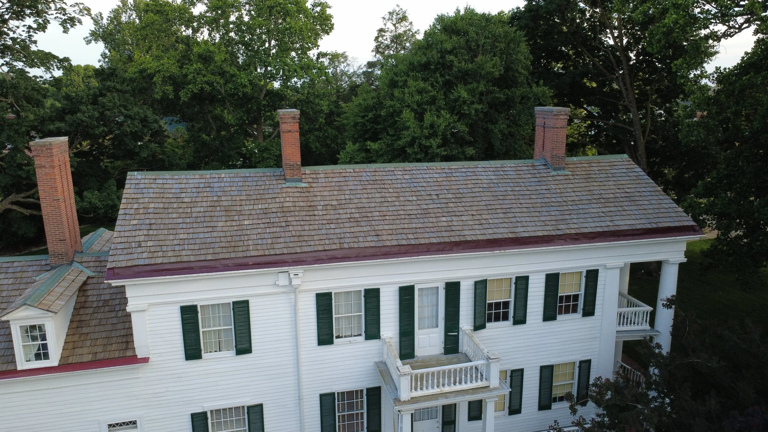 Aerial view of a large white house with green shutters, multiple stories, and a balcony, surrounded by lush green trees.
