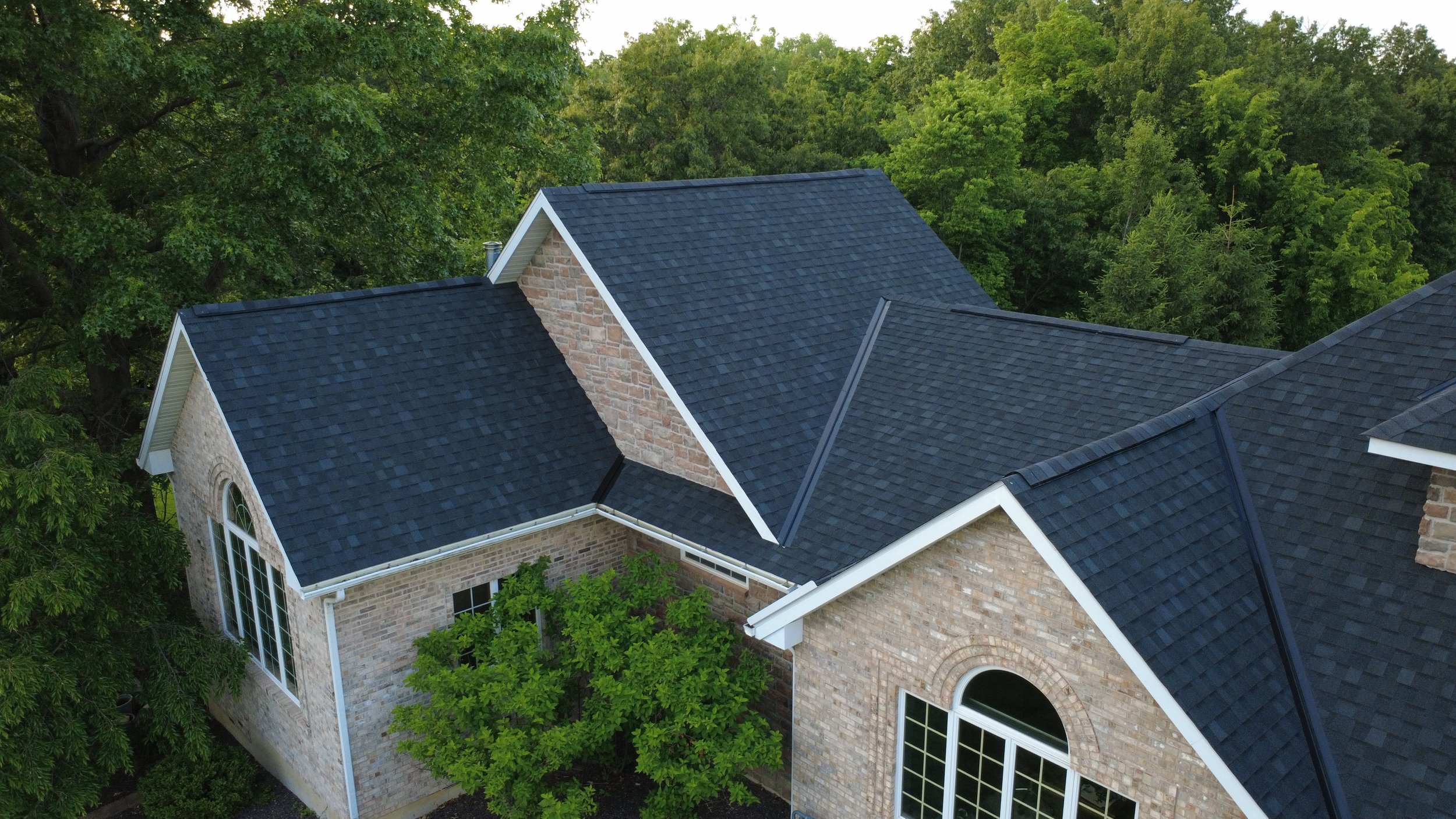 Aerial view of a house with a brick exterior and large windows, surrounded by dense green trees, with a dark blue shingle roof.