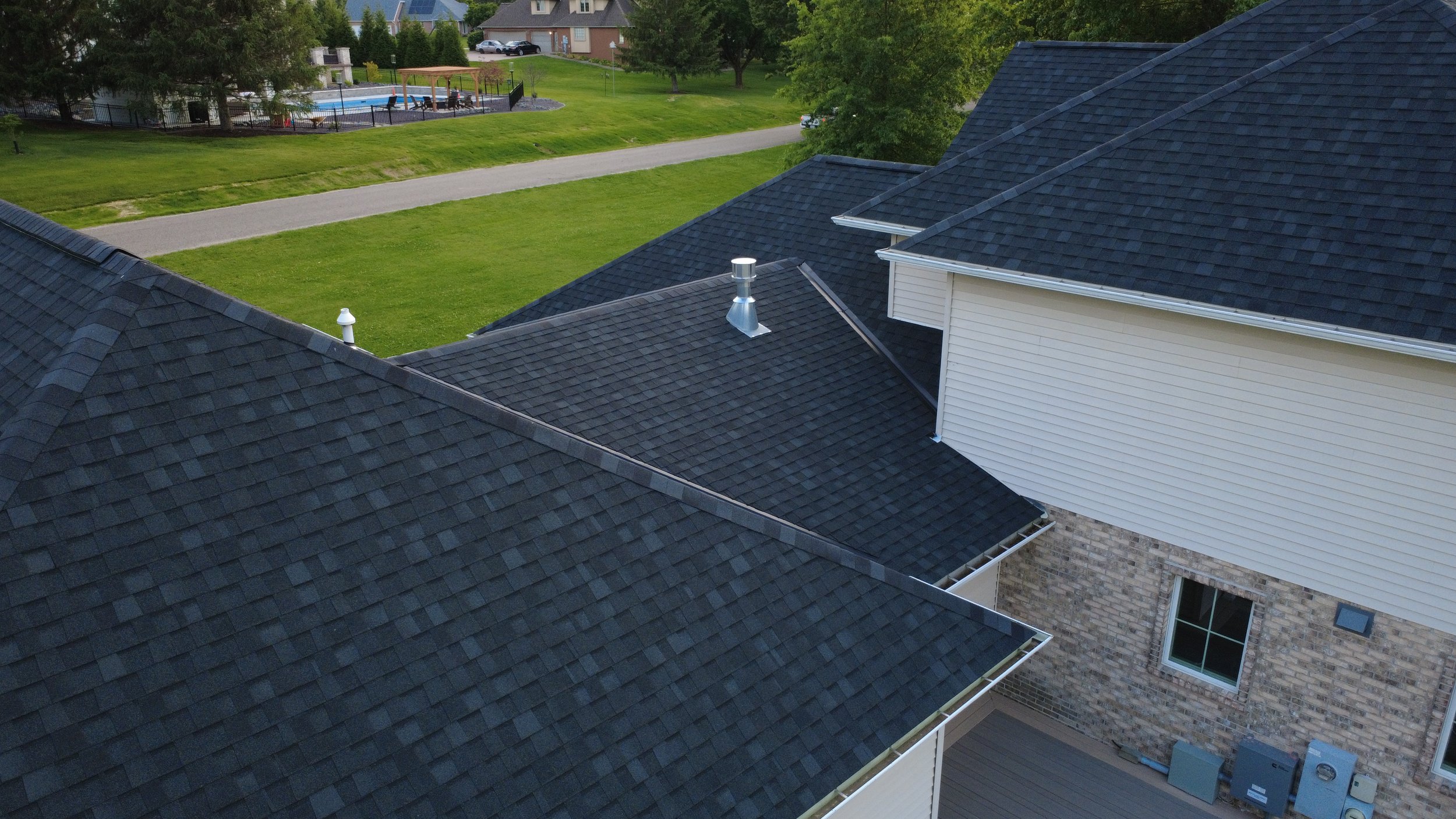 Aerial view of rooftops with dark shingle roofs, a residential house with white siding, and a backyard with a green lawn and a small pool area with a canopy.