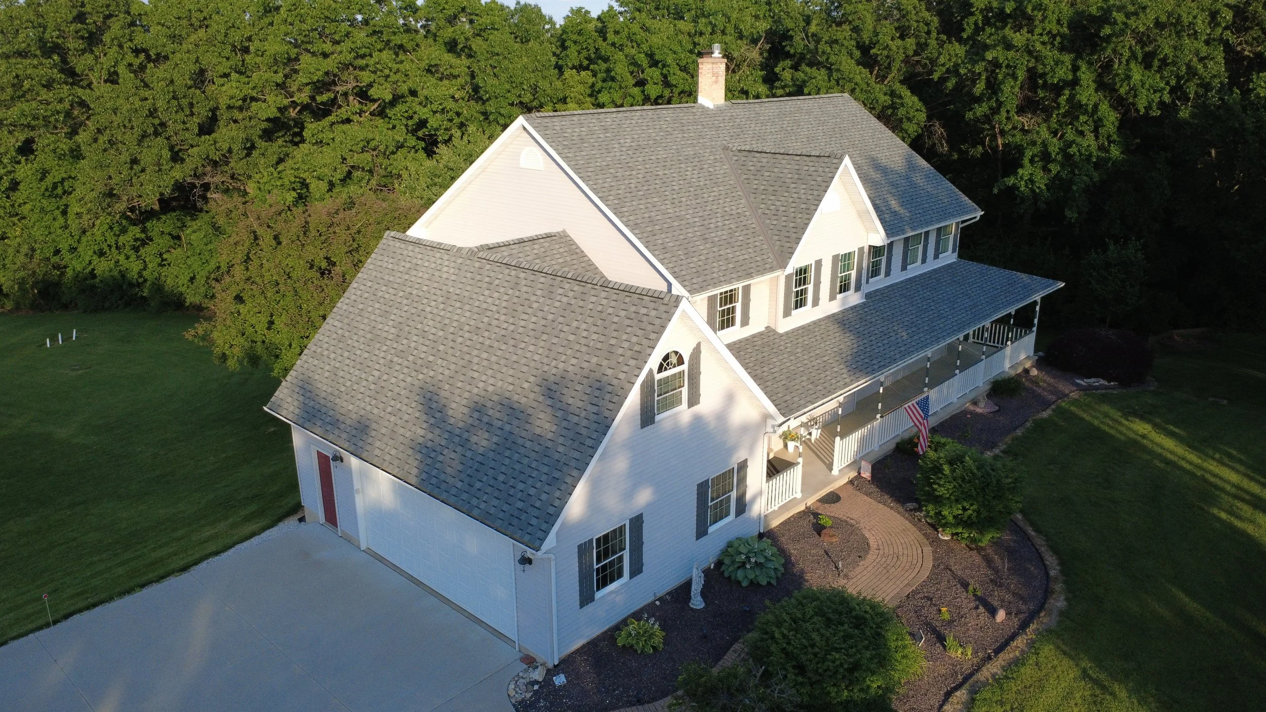 Aerial view of a two-story white house with a gray roof, surrounded by a green yard and trees, featuring a front porch with an American flag.