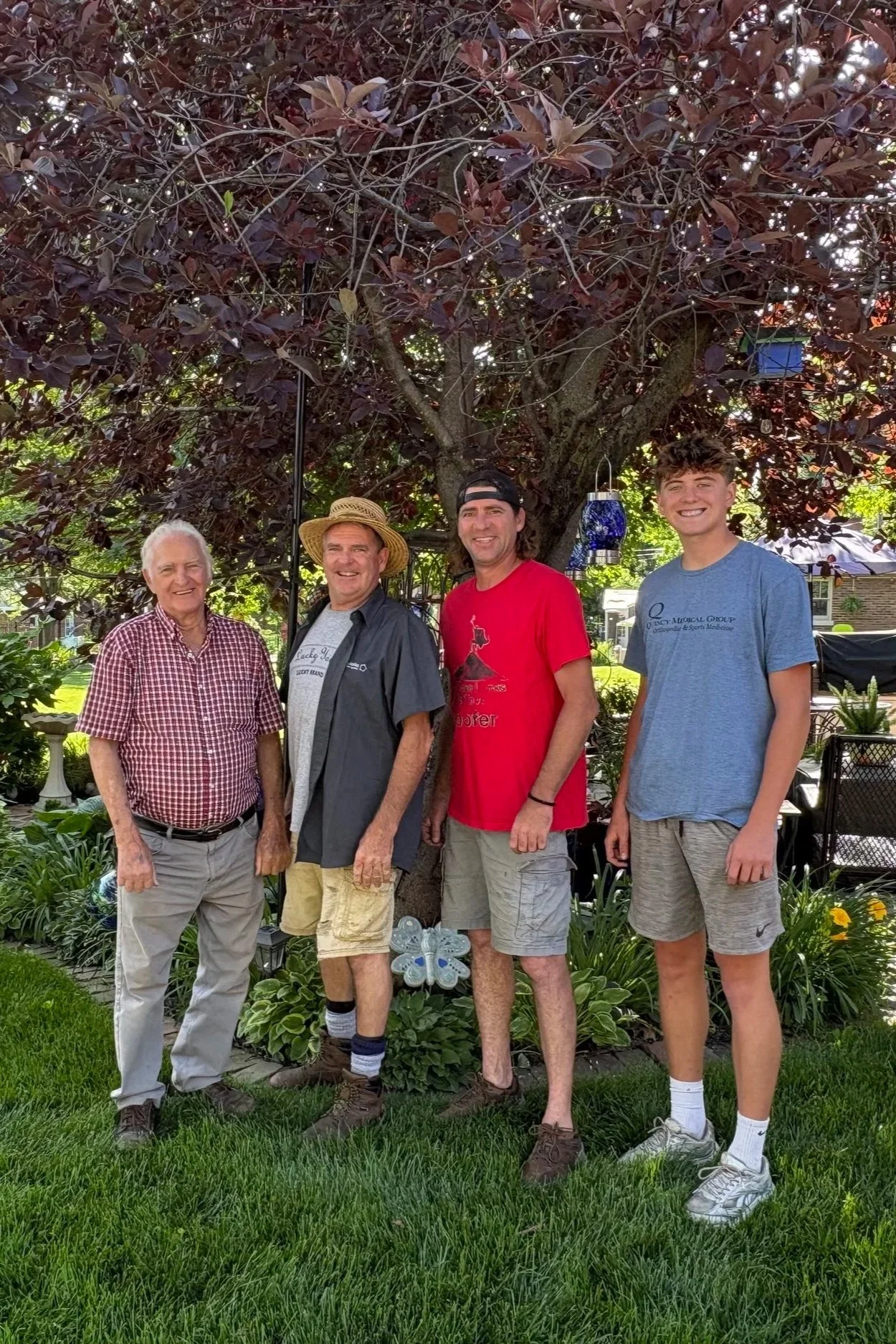 Four men standing outdoors under a tree, smiling, with garden decorations and greenery around them. They are the 4 generations of the company.