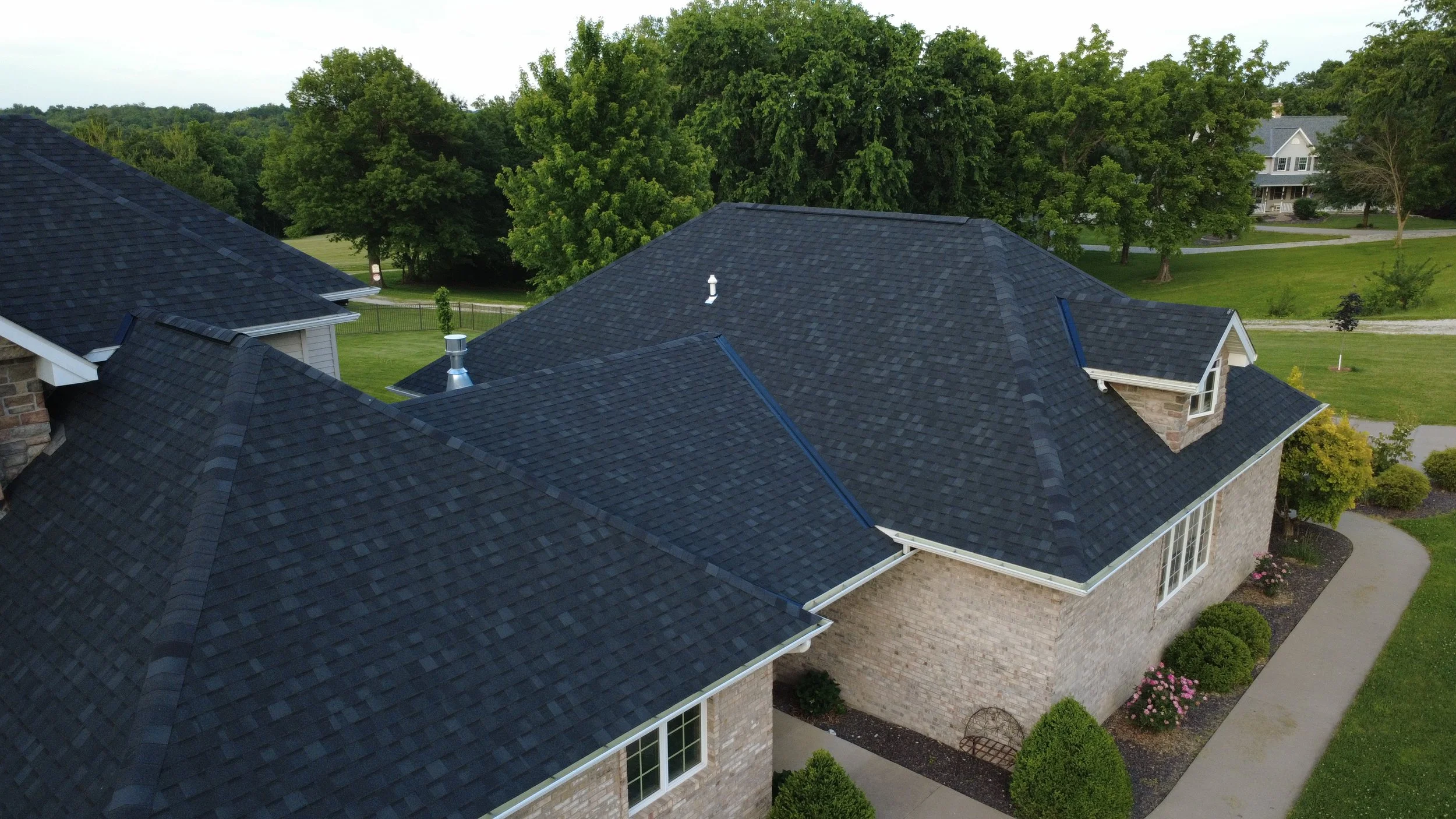 Aerial view of a house with a dark asphalt shingle roof, surrounded by green trees and a lawn.