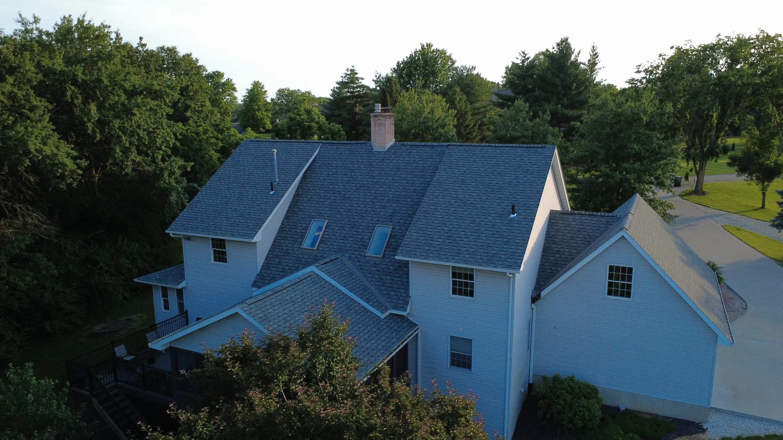 A white house with a gray shingled roof, several skylights, and a brick chimney, surrounded by green trees and a paved driveway.
