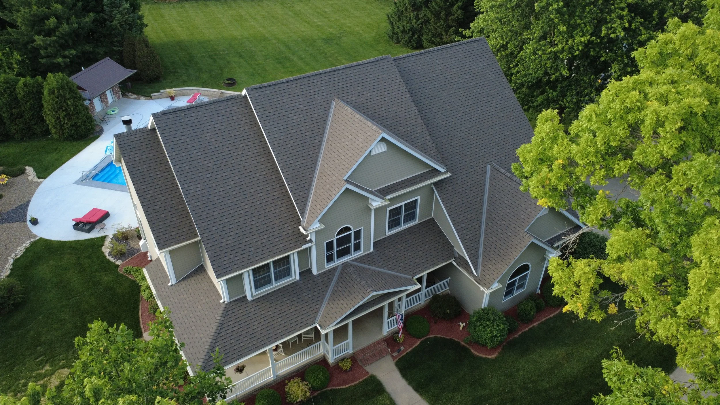 Aerial view of a large house with a tan roof, surrounded by green lawns, trees, and a backyard patio with pool and outdoor furniture.