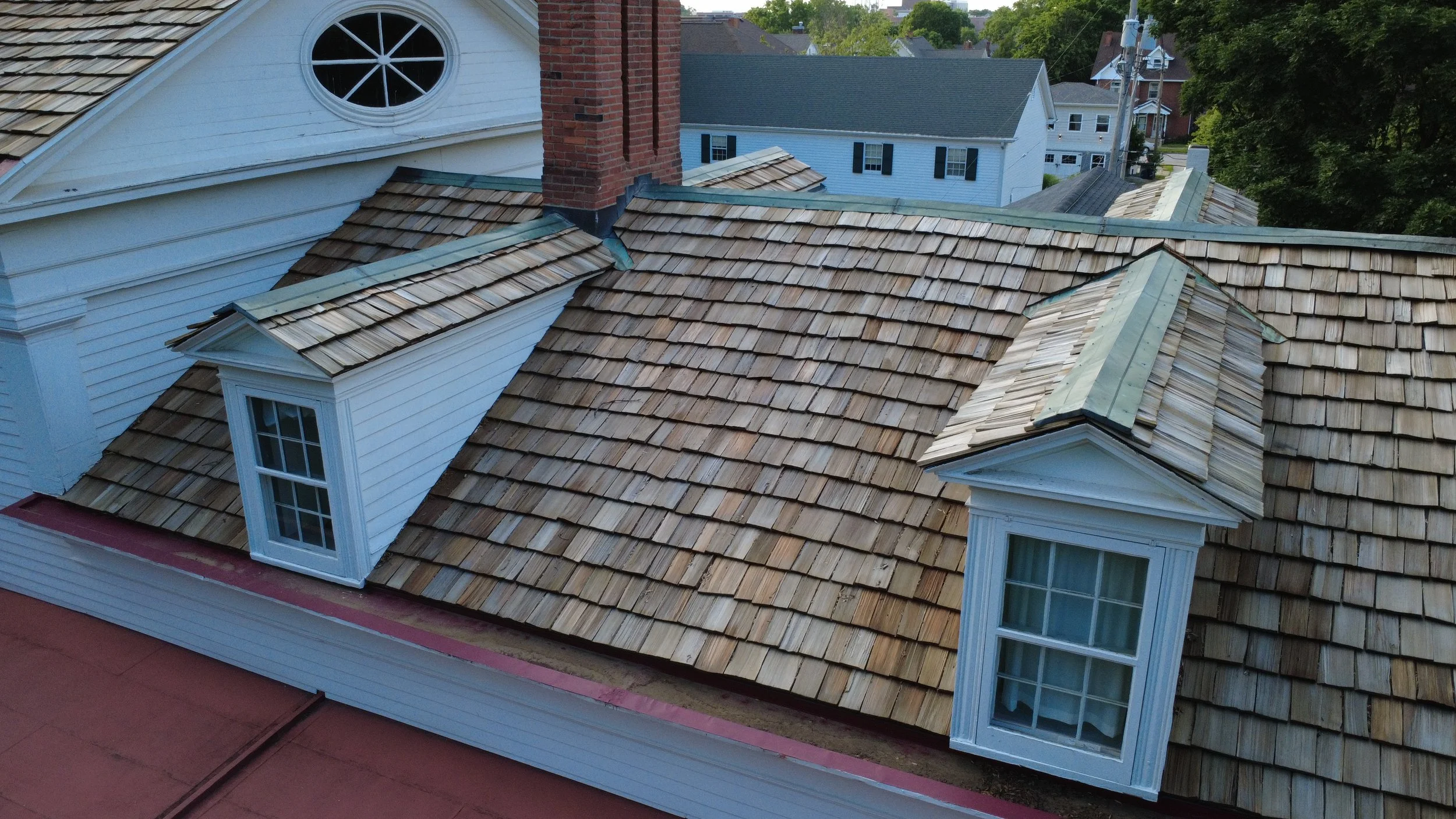 Aerial view of rooftops with wooden shingles, dormer windows, brick chimney, and neighboring houses with trees in background.