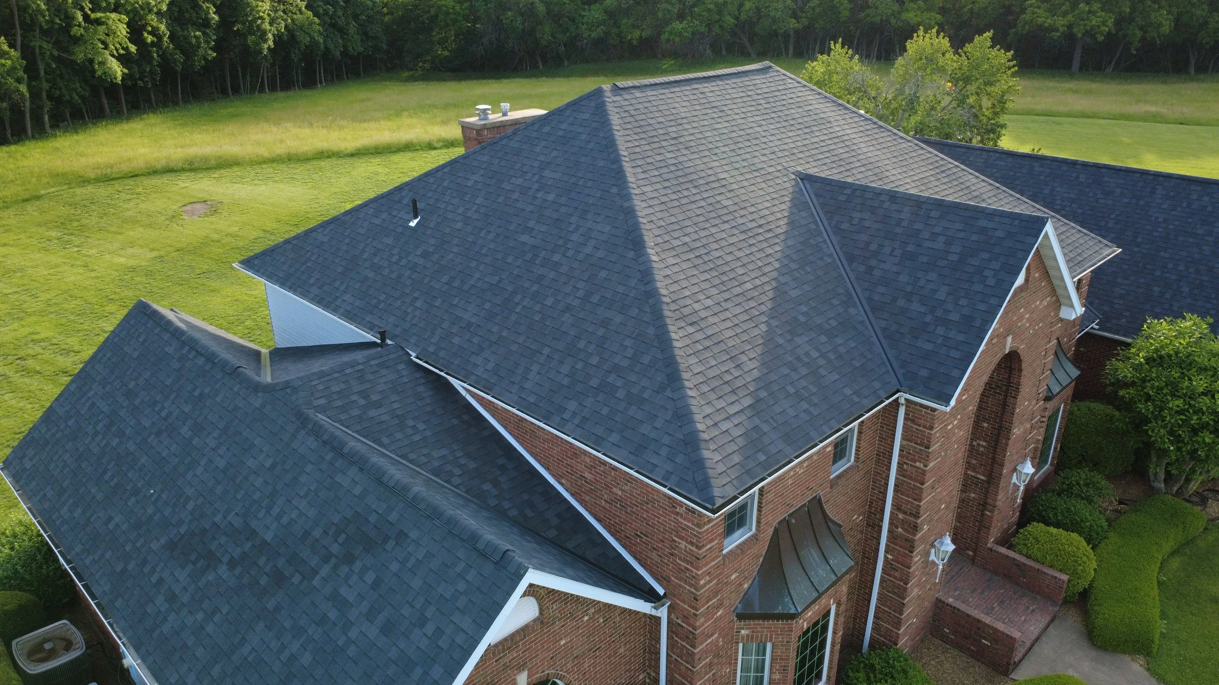 An aerial view of a house with a complex dark gray shingle roof, brick exterior walls, and a green lawn with trees surrounding the property.