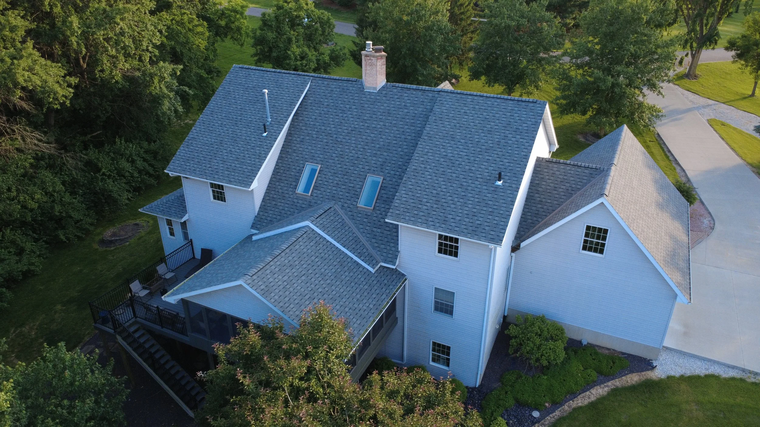 Aerial view of a large white house with a blue-grey shingled roof surrounded by trees and greenery. The house has multiple levels, skylights, and a backyard deck. There are paved pathways and a driveway visible in the yard.