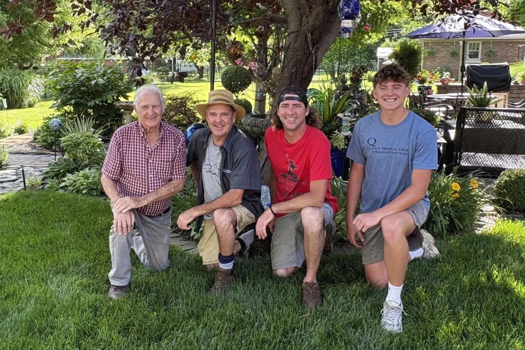 Four men kneeling and sitting outdoors in a garden with lush green grass, plants, trees, and outdoor furniture in the background. They are smiling and looking at the camera. They are the 4 generations of the family buisness.