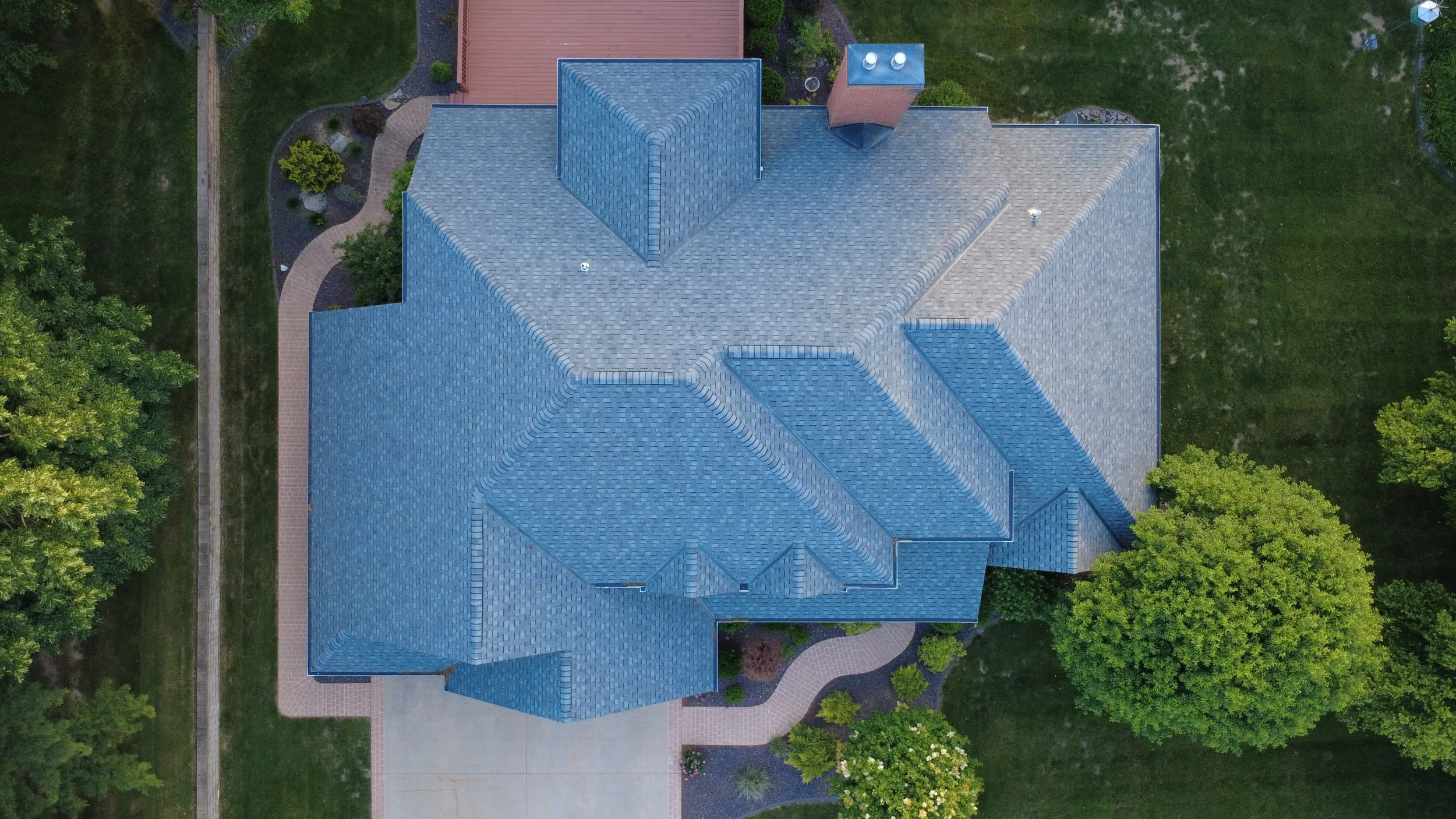 An aerial view of a house with a blue roof, paved walkways, green lawns, trees, and some garden features.