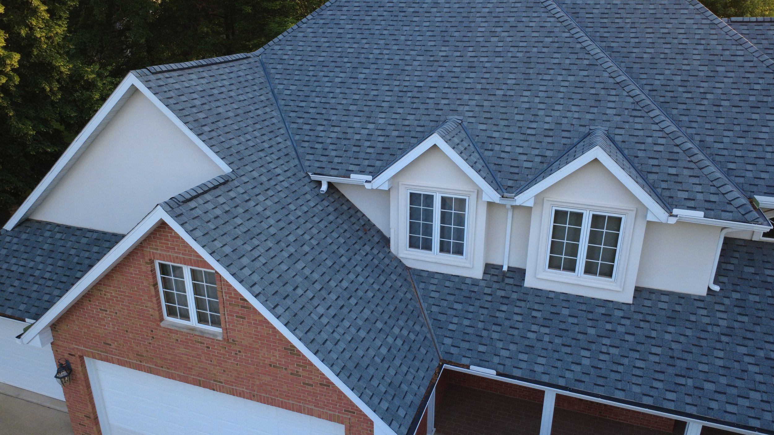 Aerial view of a house with a gray shingle roof, white dormer windows, and a red brick exterior, surrounded by trees.