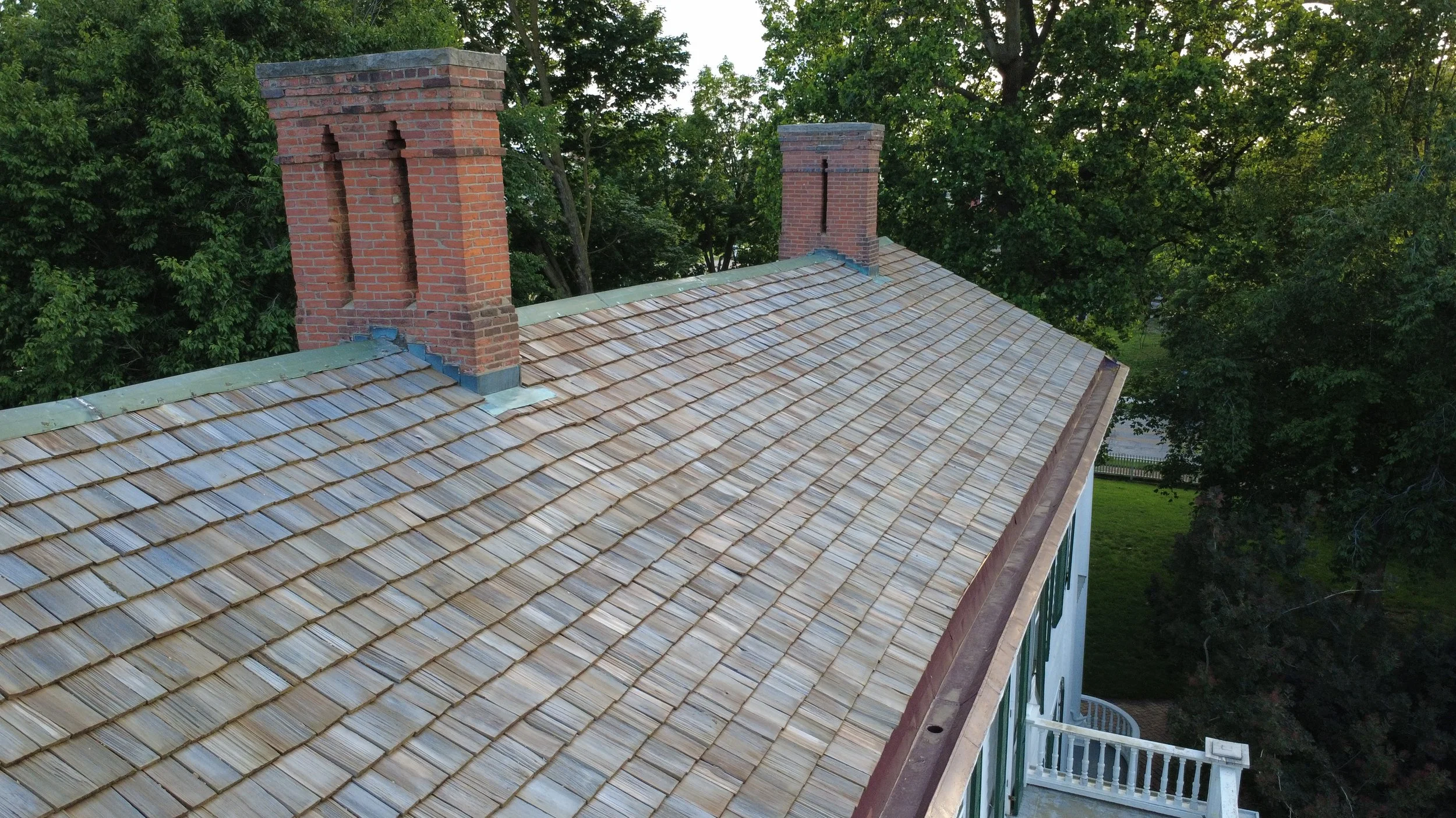 View of a rooftop with tan shingles, accented by three red brick chimneys, surrounded by green trees.