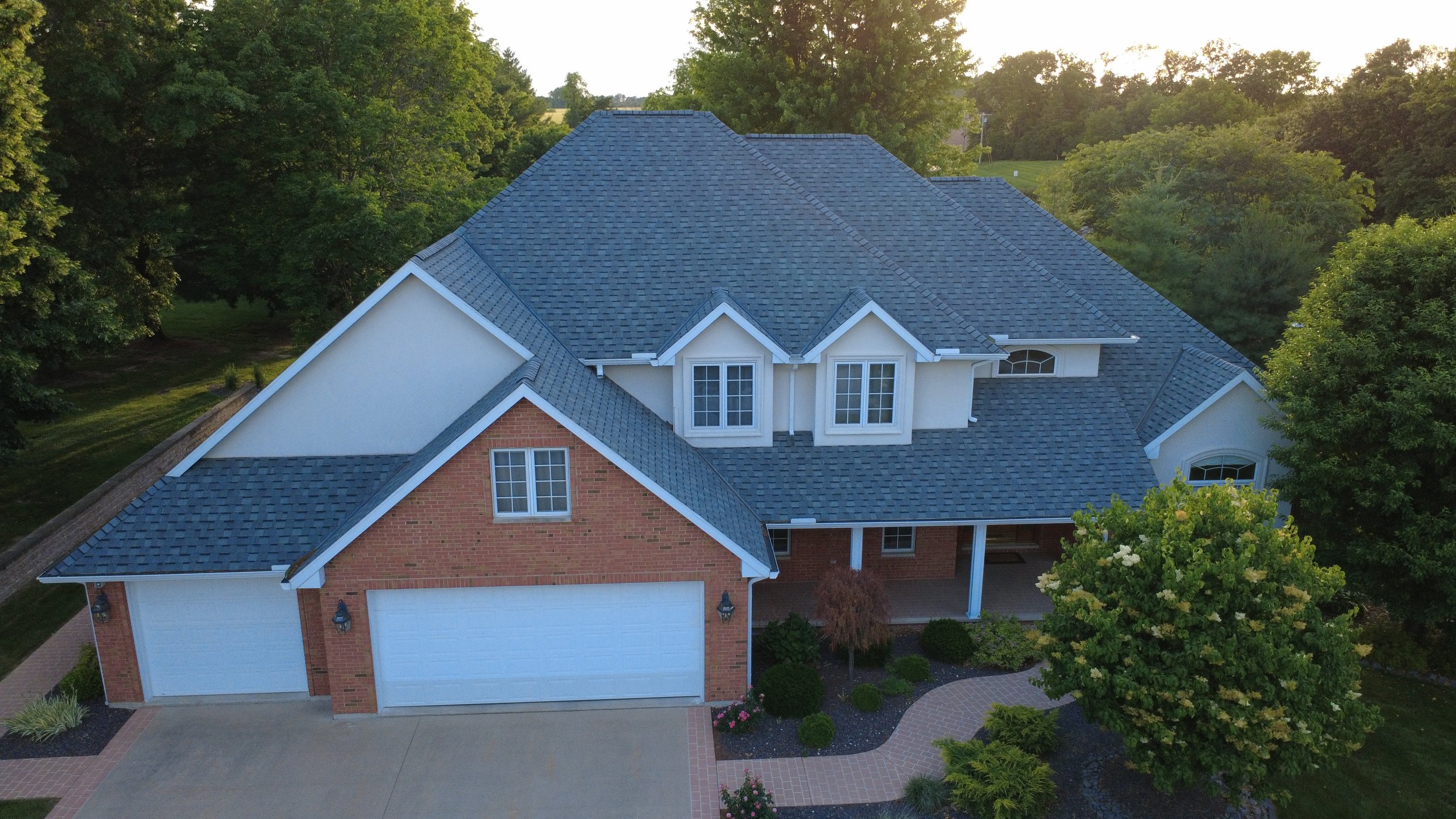 A large house with a blue shingled roof, white walls, and a brick garage door, surrounded by green trees and a well-maintained yard.