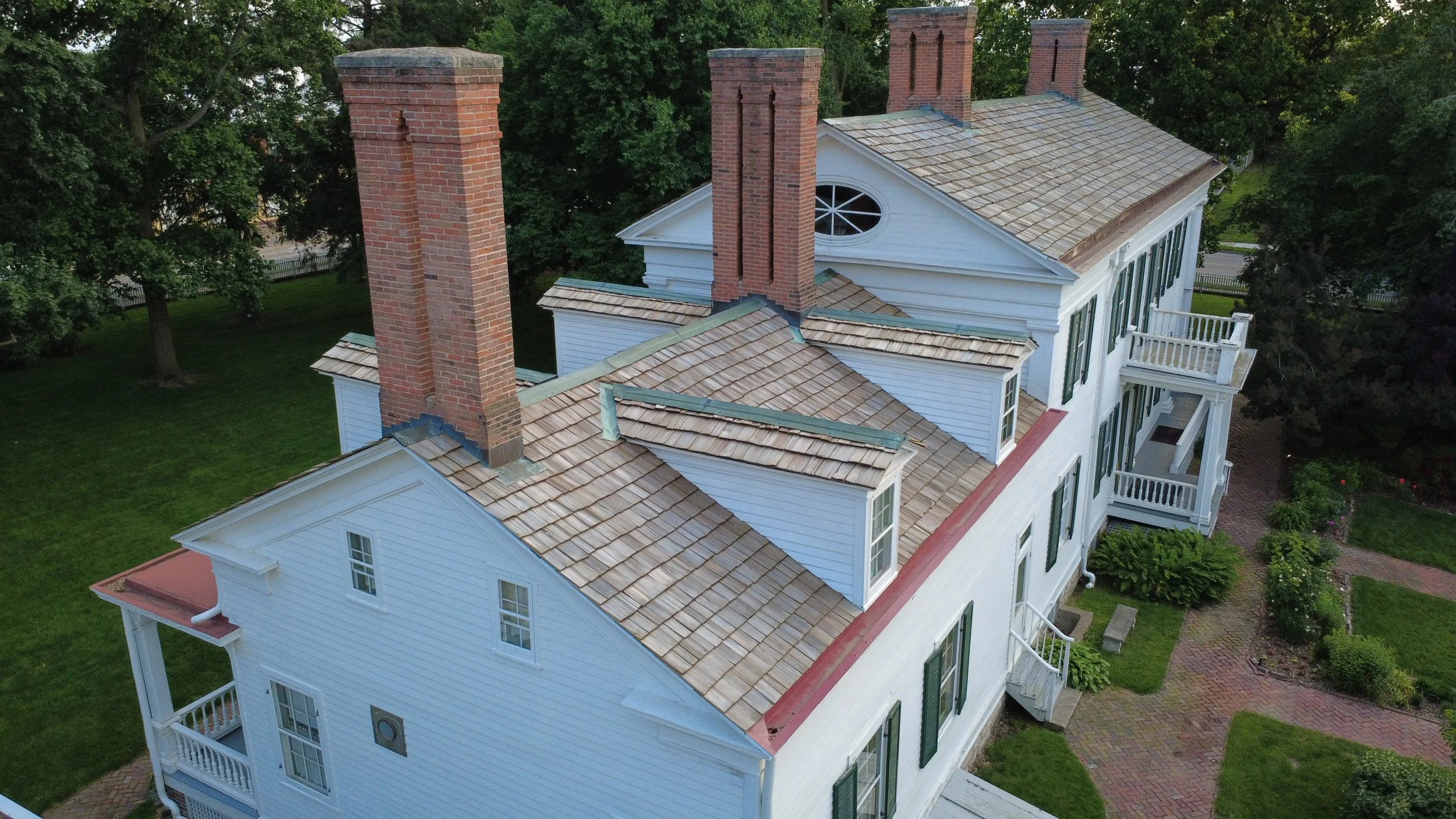 Aerial view of a large white house with multiple chimneys and a gabled roof, surrounded by green trees and a brick walkway.