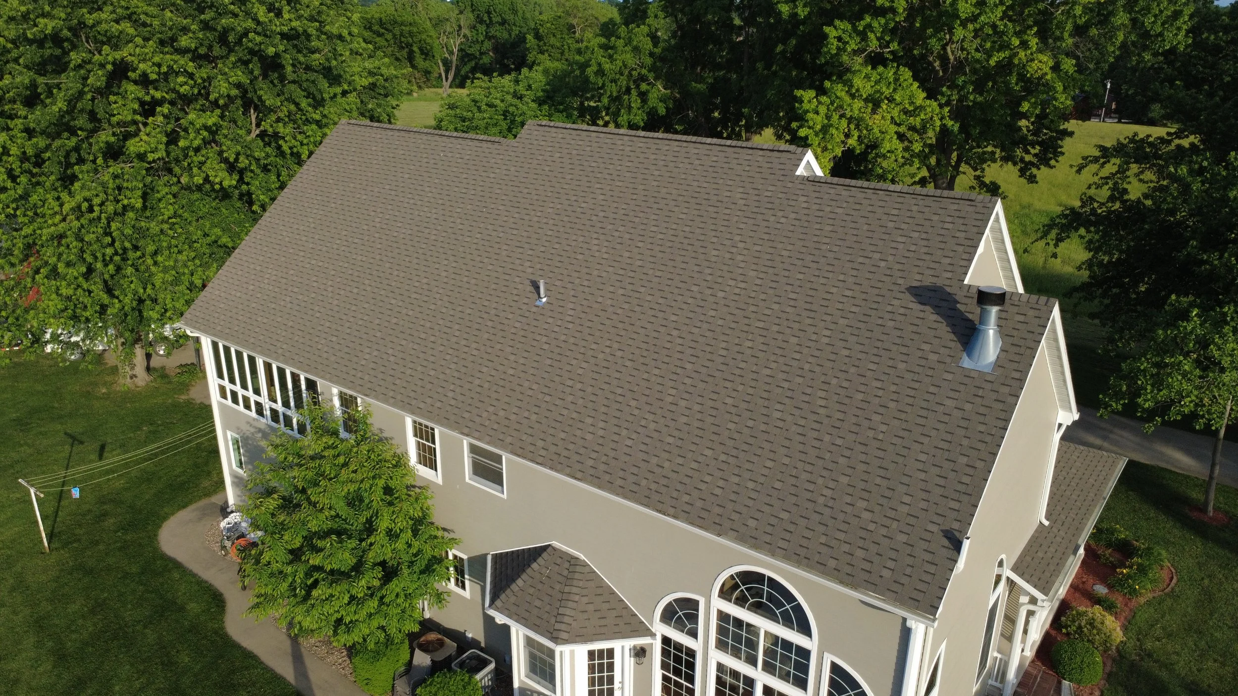 Aerial view of a large house with a gray shingle roof surrounded by green trees and grass.