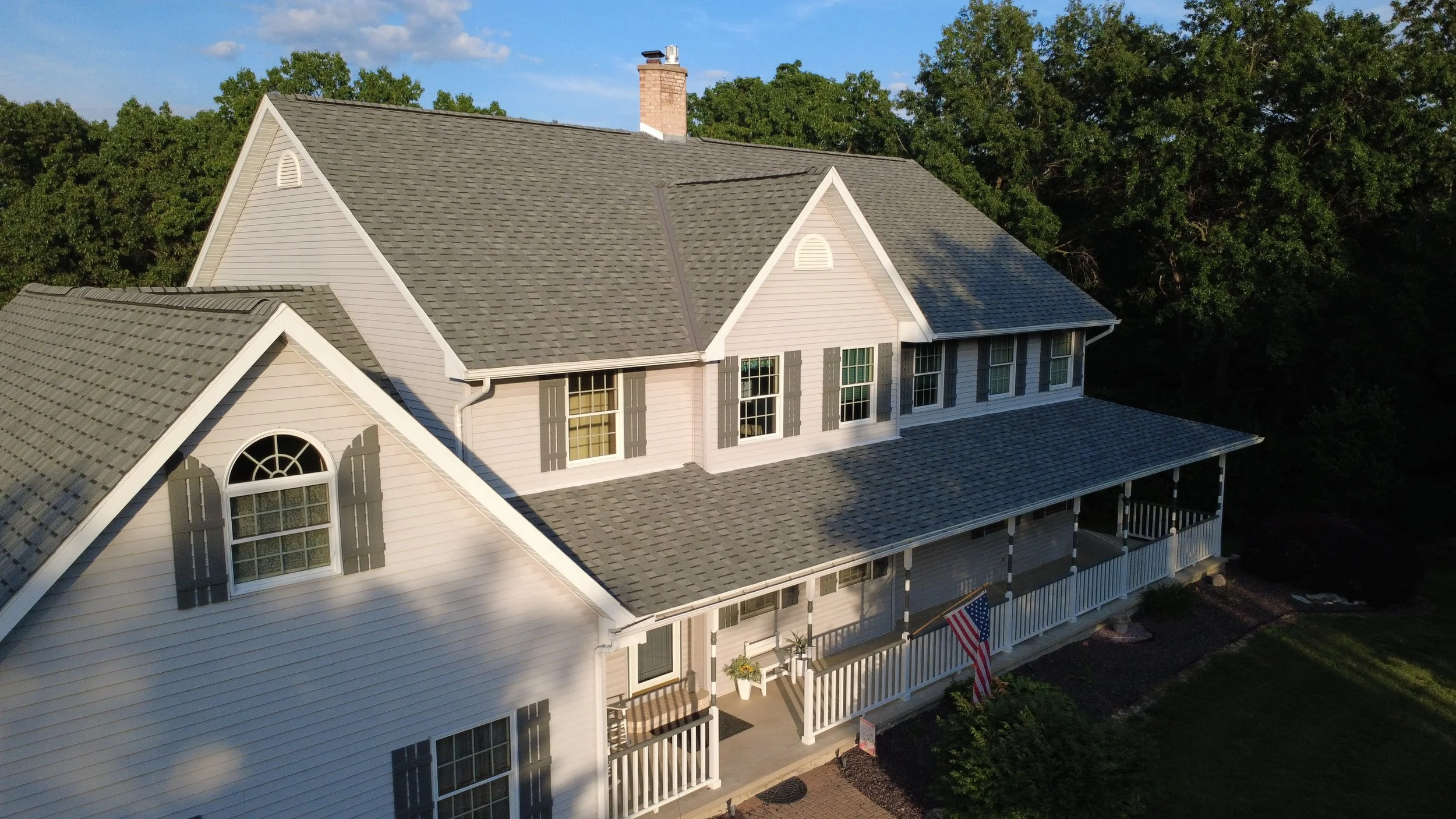 Aerial view of a large light-colored house with multiple gable roofs, a porch with white railing, and a decorated American flag hanging outside, surrounded by green trees.