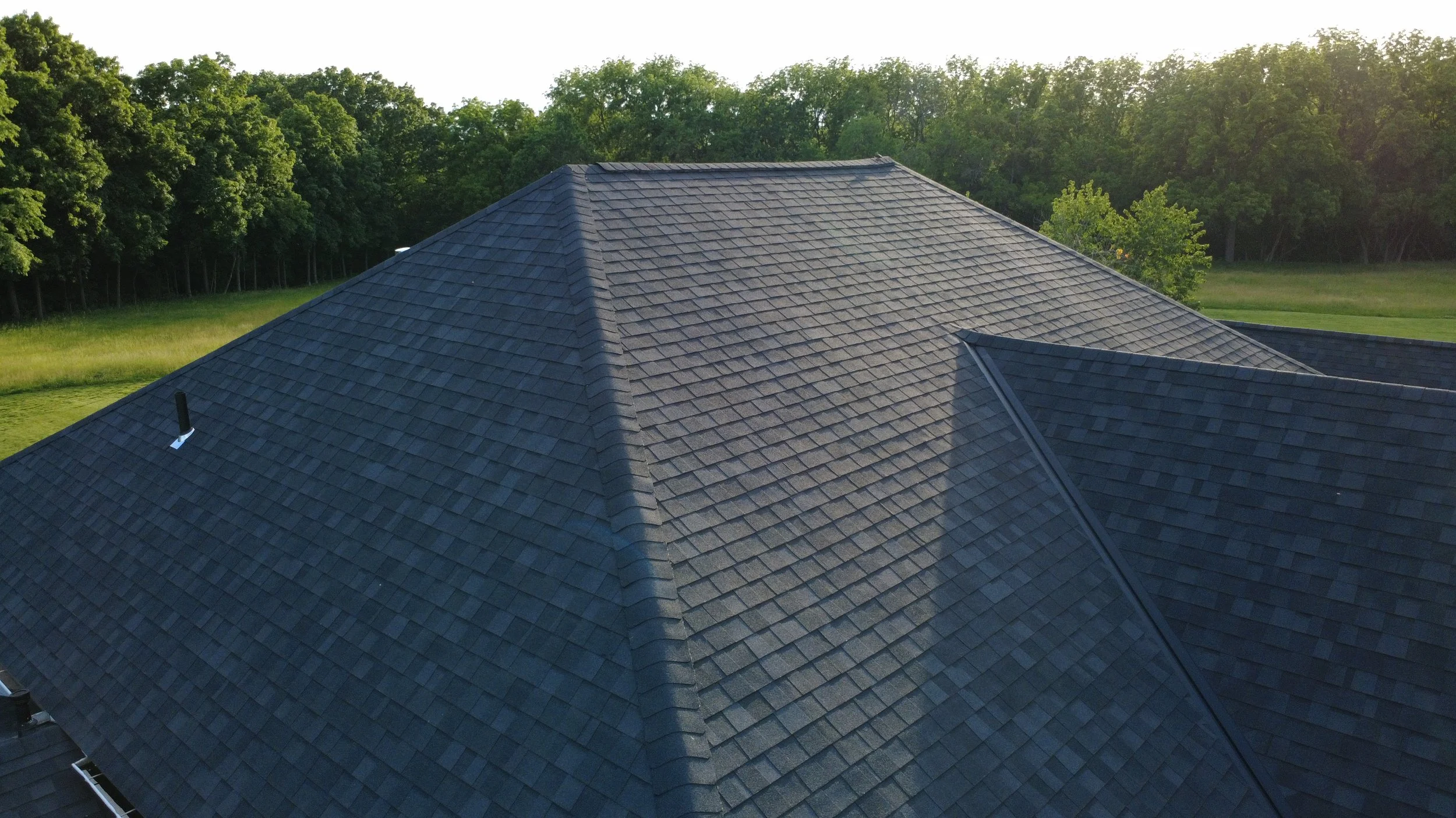 A house roof with dark-colored shingles surrounded by green trees and grassy fields, with sunlight in the background.