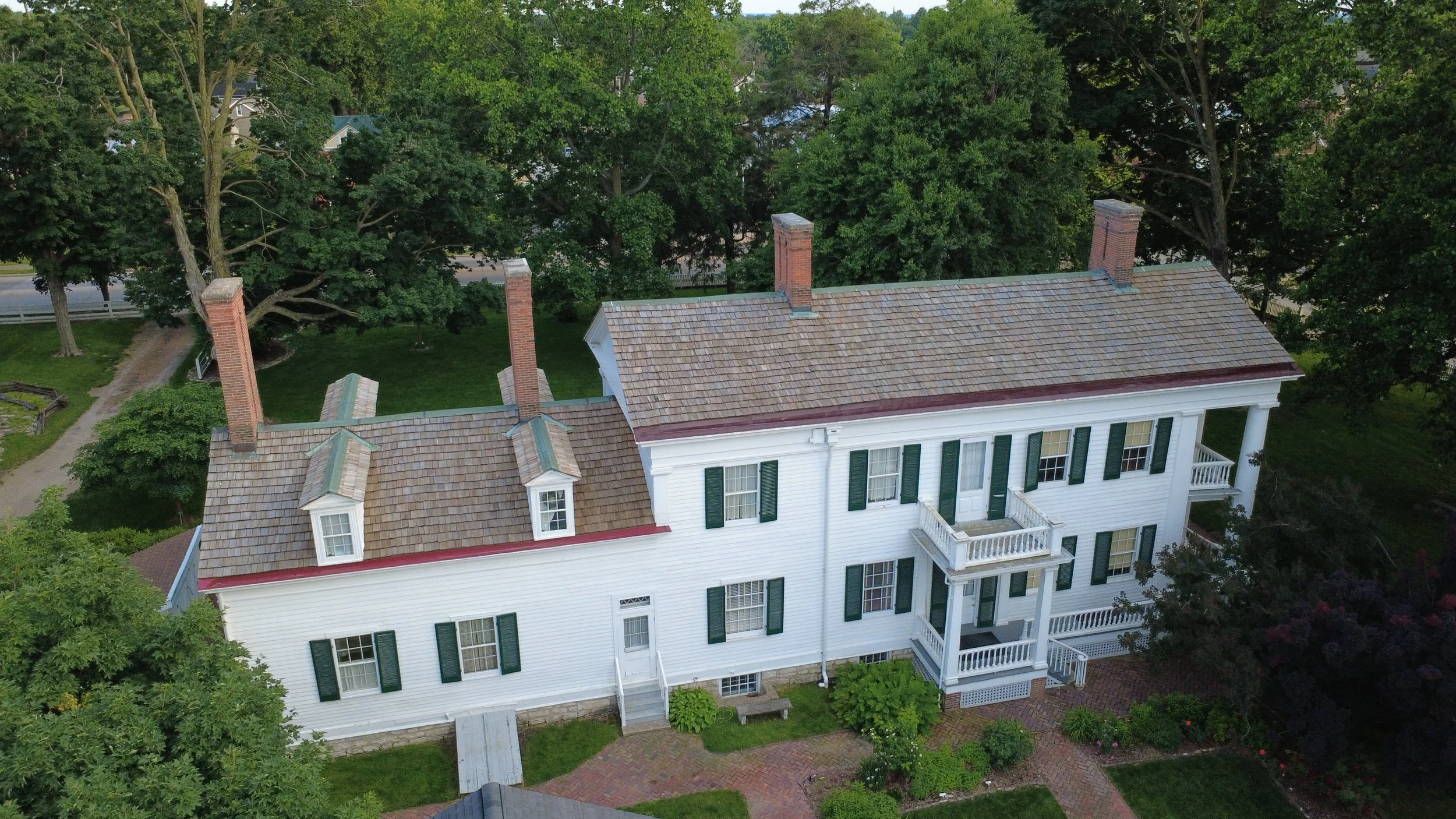 Aerial view of a large white house with a brown shingle roof and green shutters, surrounded by green trees and a brick pathway in a yard.