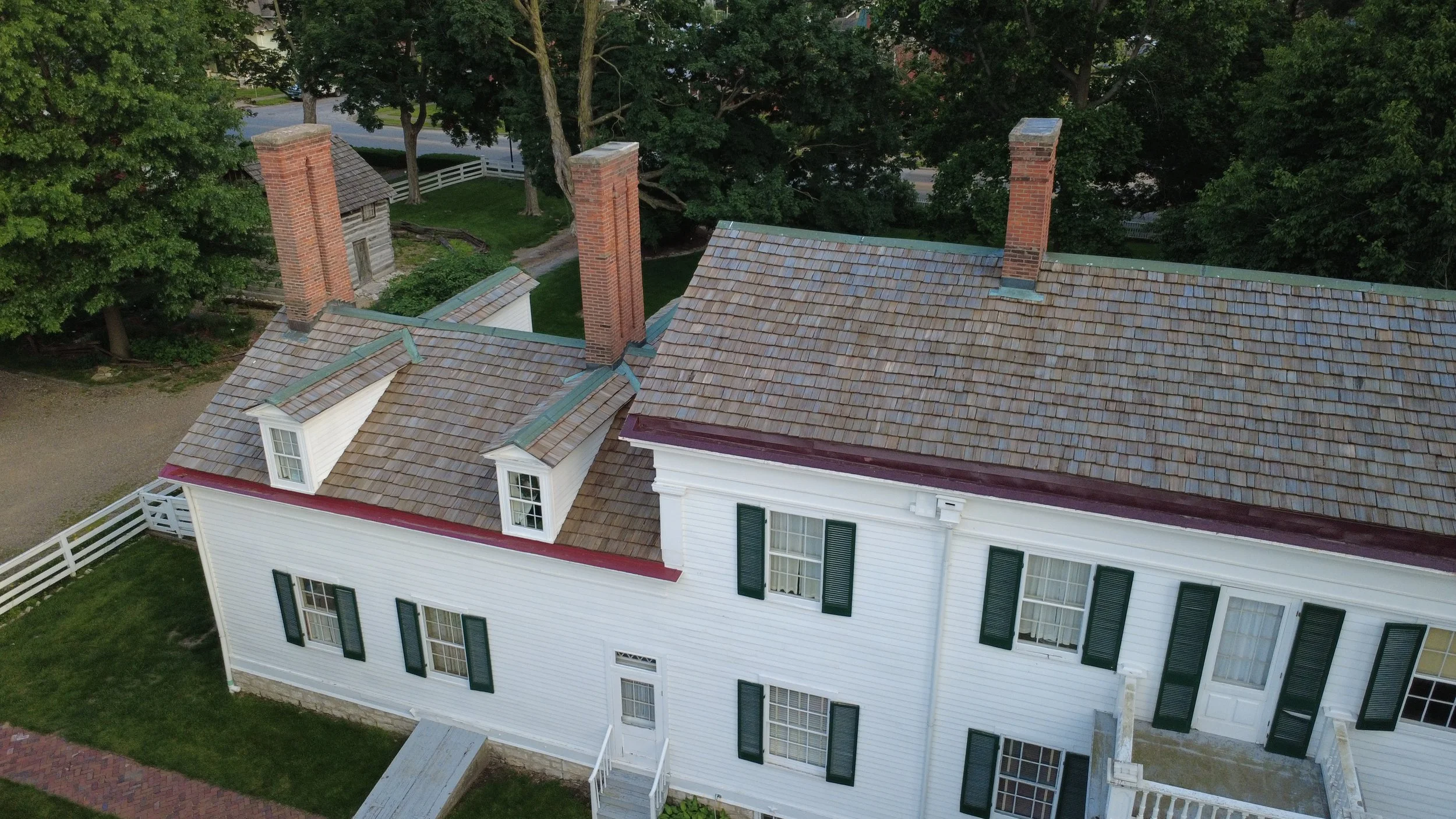 Aerial view of a white house with black shutters, multiple brick chimneys, copper and slate roof sections, surrounded by trees and a white fence.