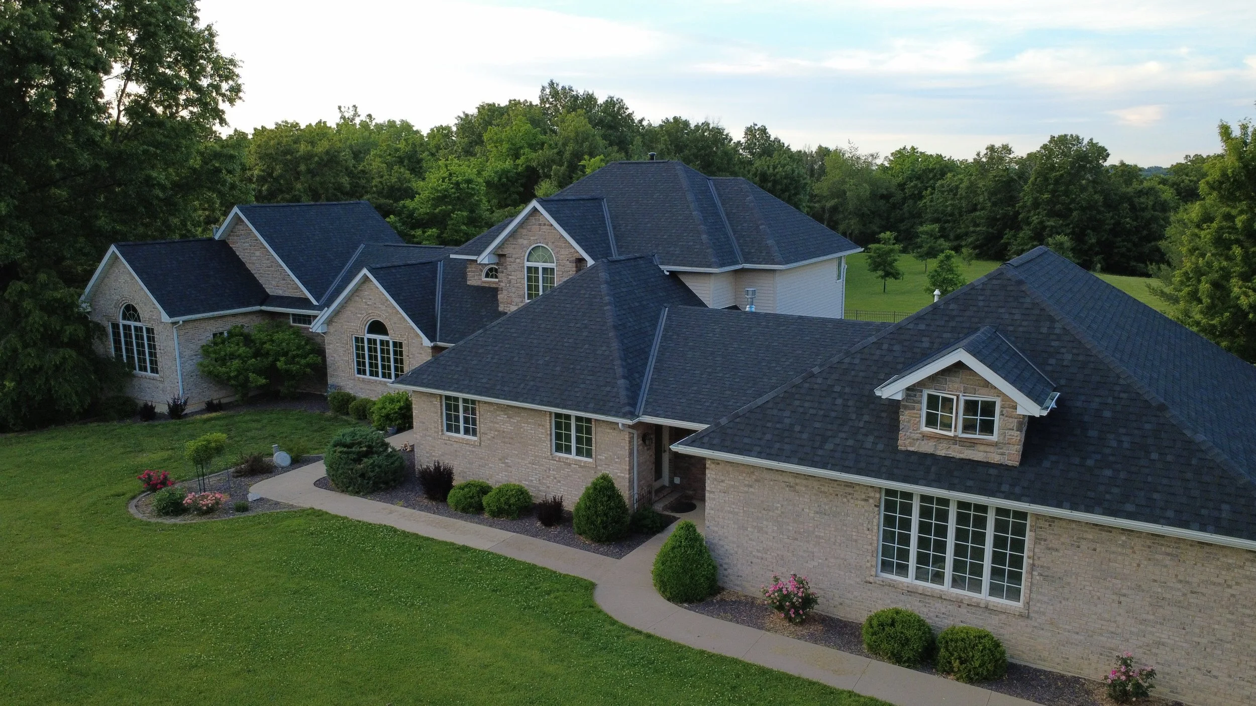 A large, multi-story brick house with a dark shingled roof, surrounded by well-manicured lawn and landscaping, with trees and greenery in the background.