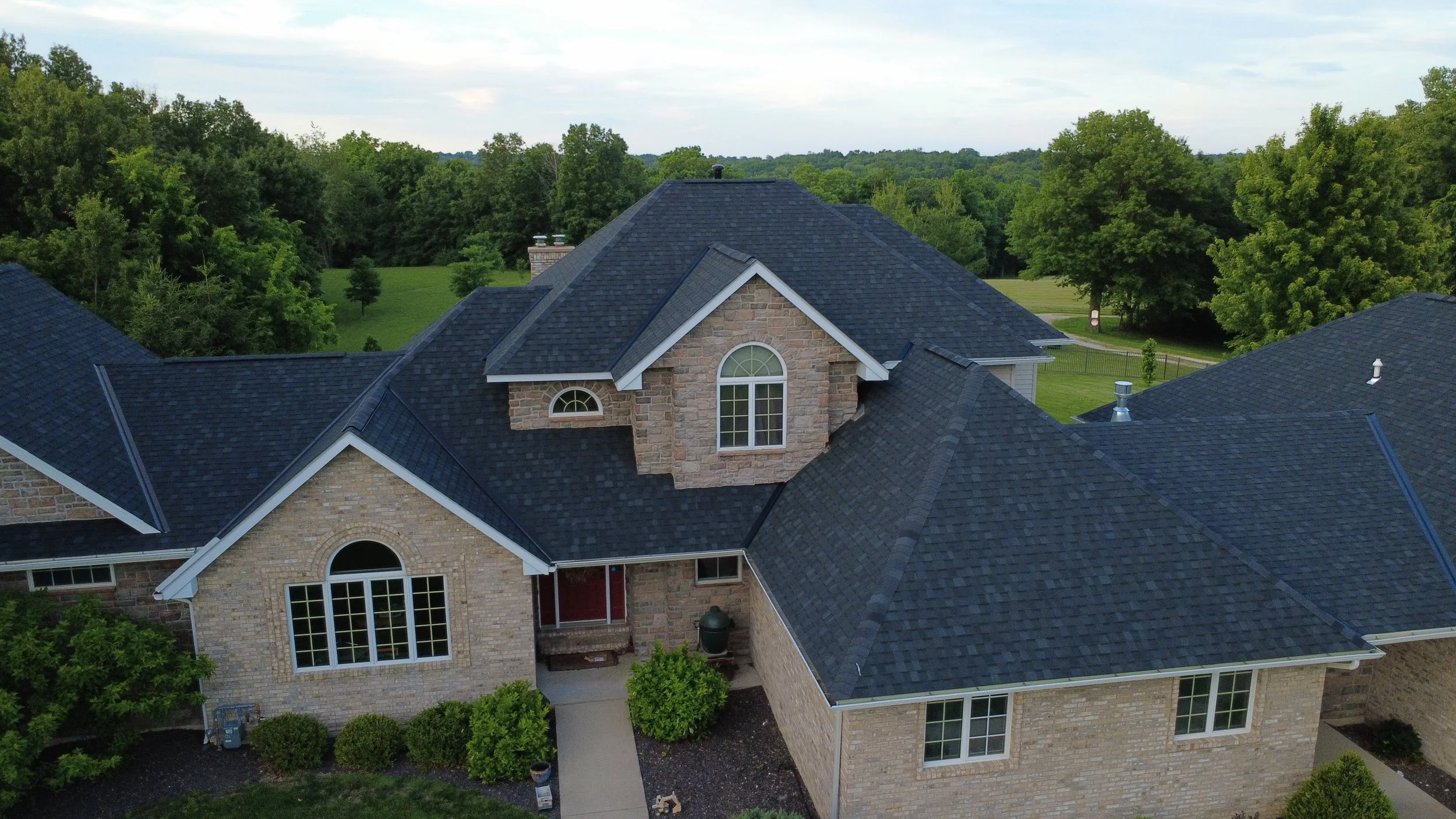 Aerial view of a house with brick walls, multiple gable roofs with dark shingles, large arched and rectangular windows, surrounded by green trees and a lawn.