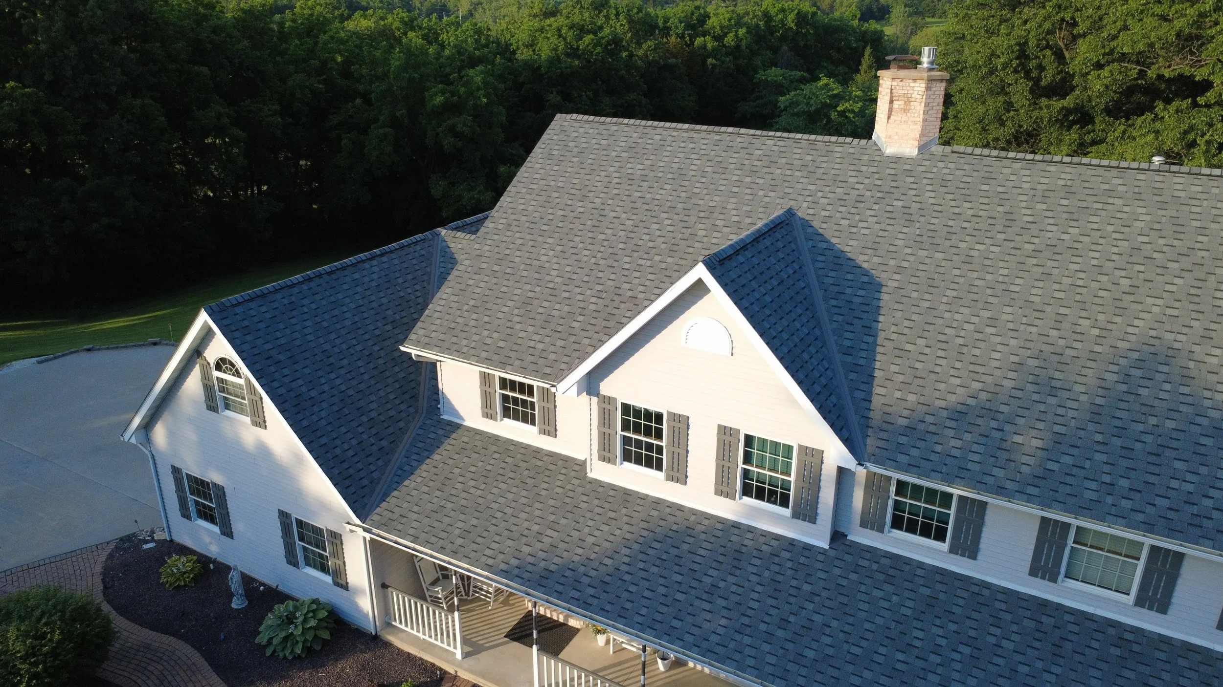 Aerial view of a large white house with a gray shingled roof and multiple gables, surrounded by trees and green landscape.