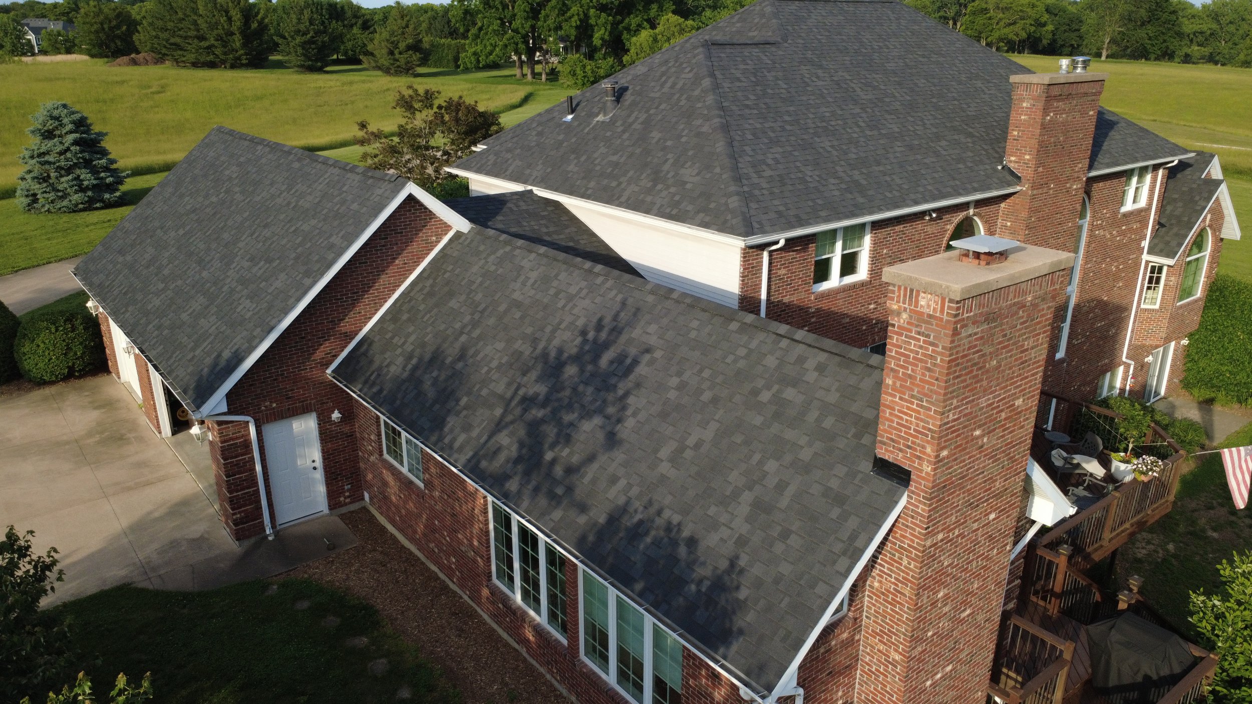 Aerial view of a brick house with multiple gable roofs, a large brick chimney, surrounded by green lawns and trees.
