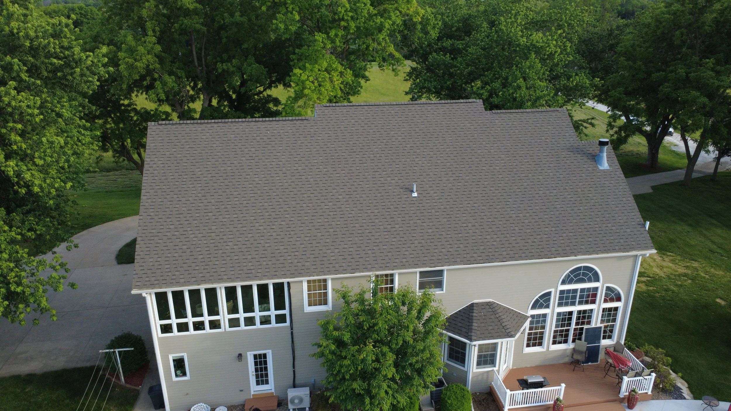 Aerial view of a two-story house with a gray shingle roof, surrounded by green trees and a lawn, with a concrete driveway and a backyard patio with outdoor furniture.
