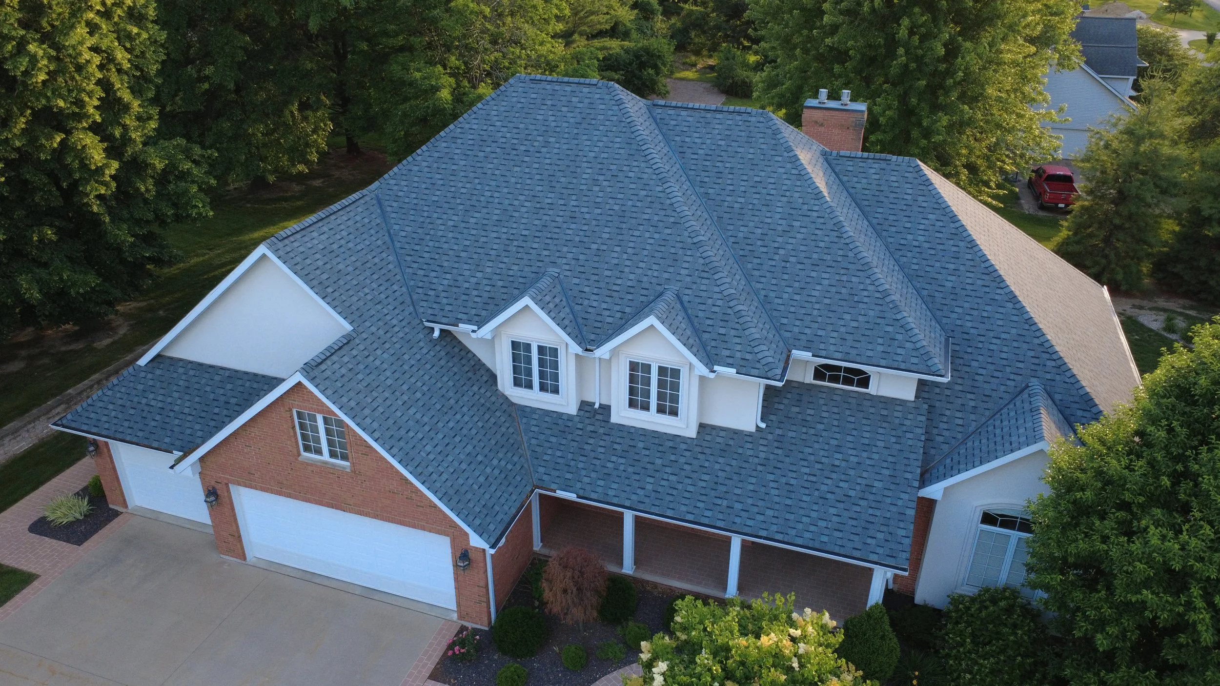 An aerial view of a house with a complex blue shingled roof, brick and white siding, surrounded by trees and greenery.