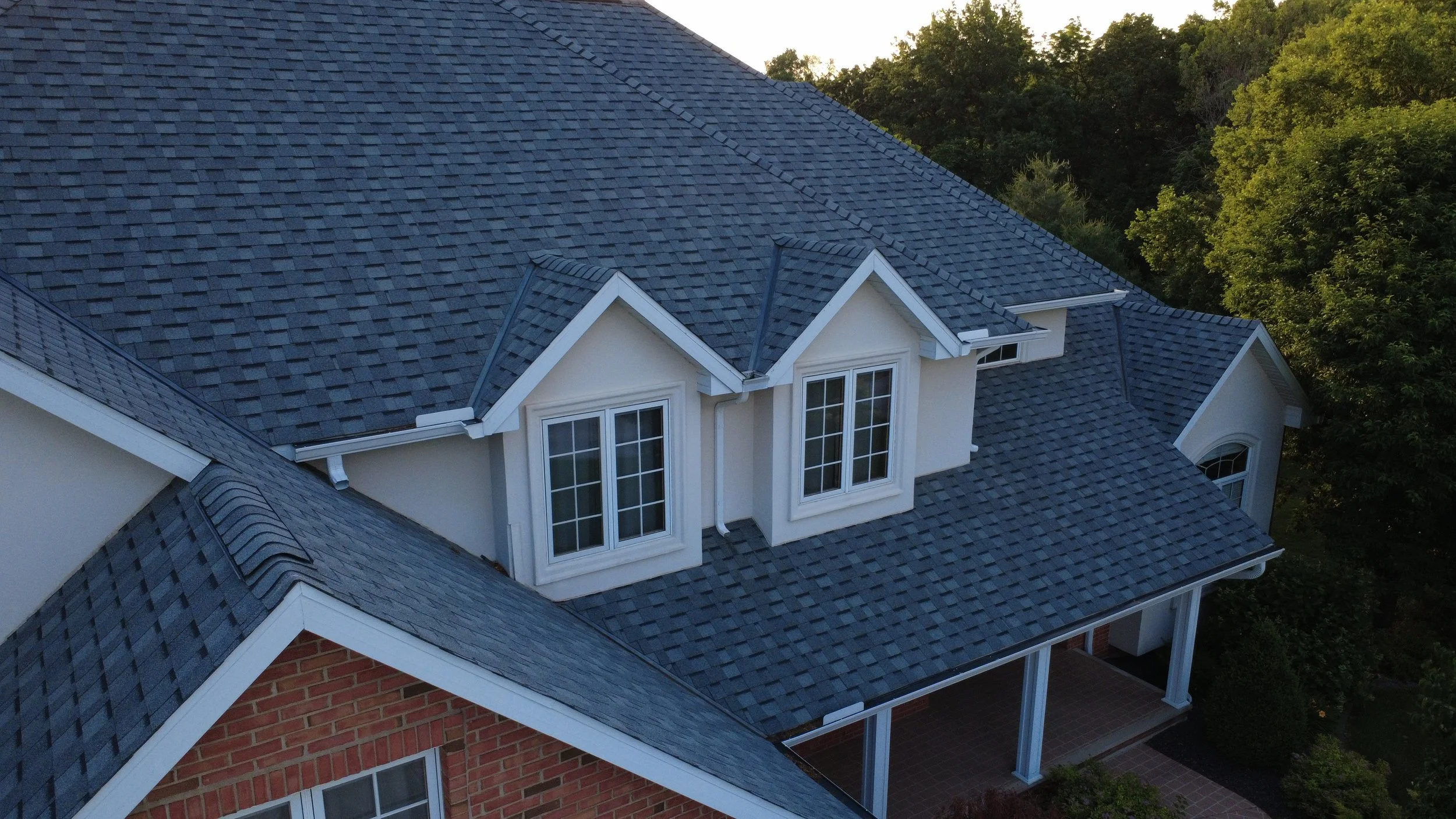 Aerial view of a house with gray shingle roof, dormer windows, and a combination of brick and white siding, surrounded by green trees.