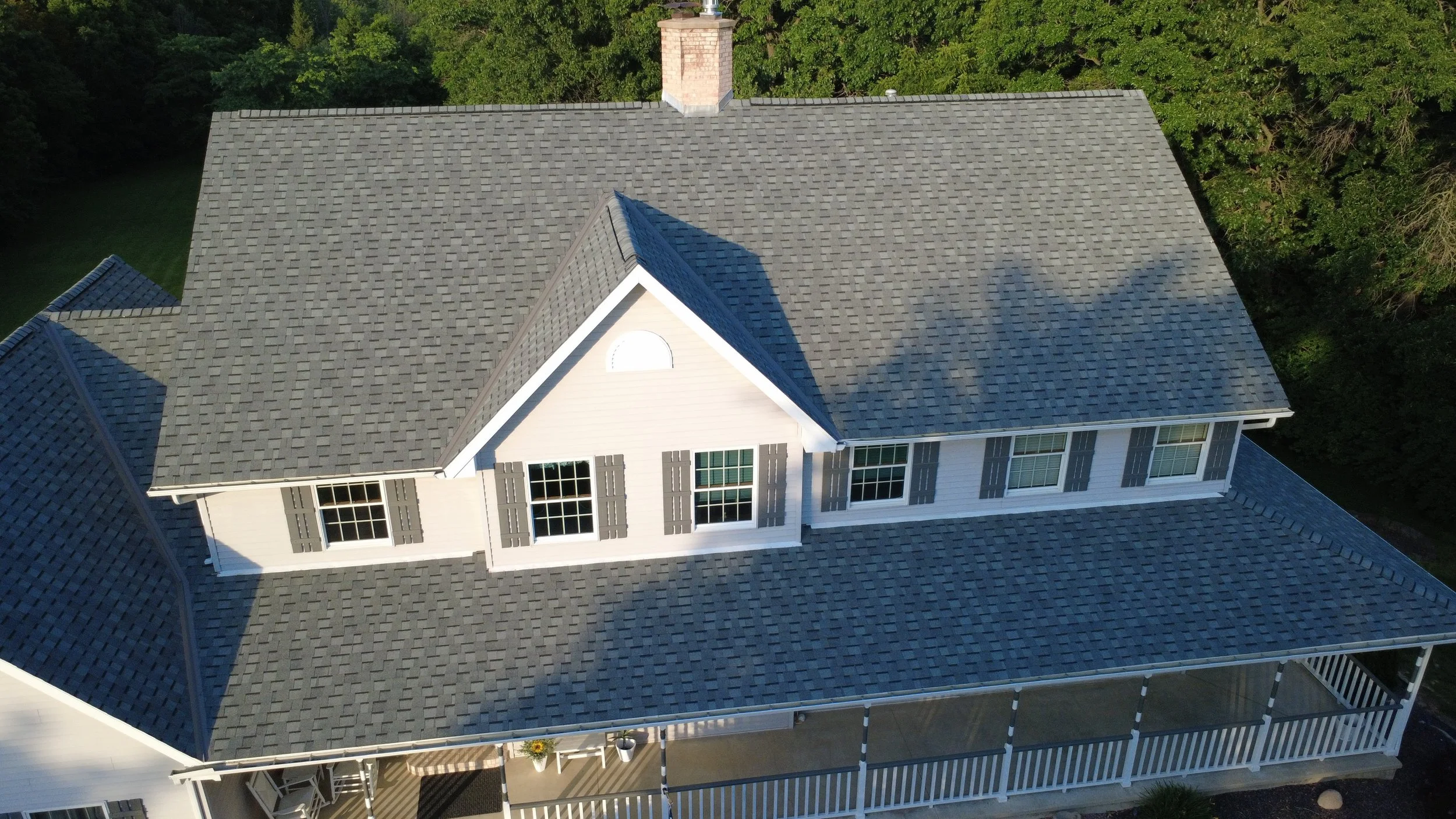 Aerial view of a house with a gray shingle roof, white exterior walls, multiple windows with gray shutters, and a large porch with white railings. Surrounding trees are visible in the background.